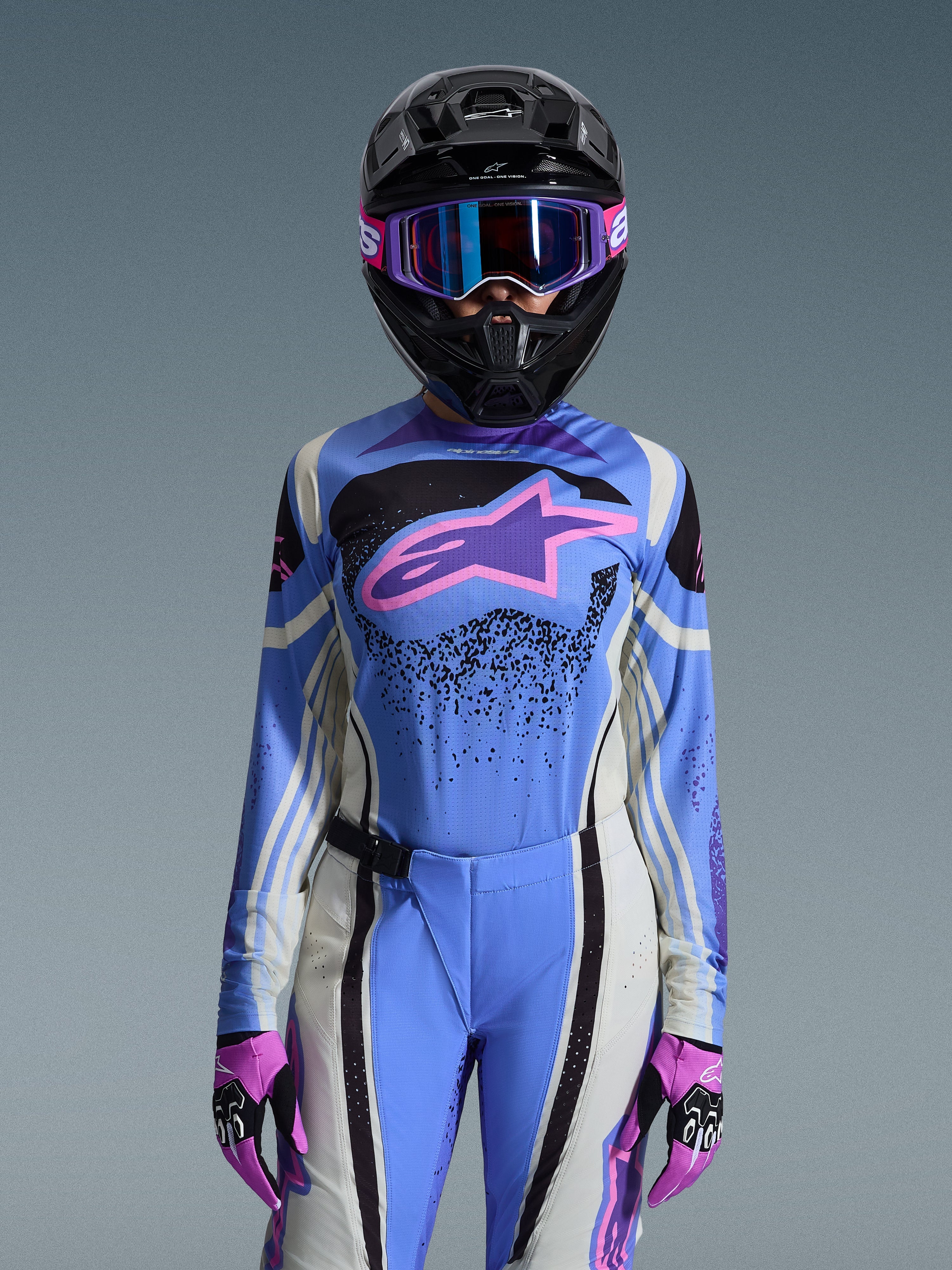 Kvindelig motocrosskører iført Alpinestars 2026 Stella Techstar Nomur-udstyrssæt, blå jersey med lyserødt og lilla stjernelogo, matchende blå bukser med hvide og sorte sidepaneler, sort hjelm med lilla og blå goggles, lyserøde og sorte handsker, komplet off-road racersæt til kvinder