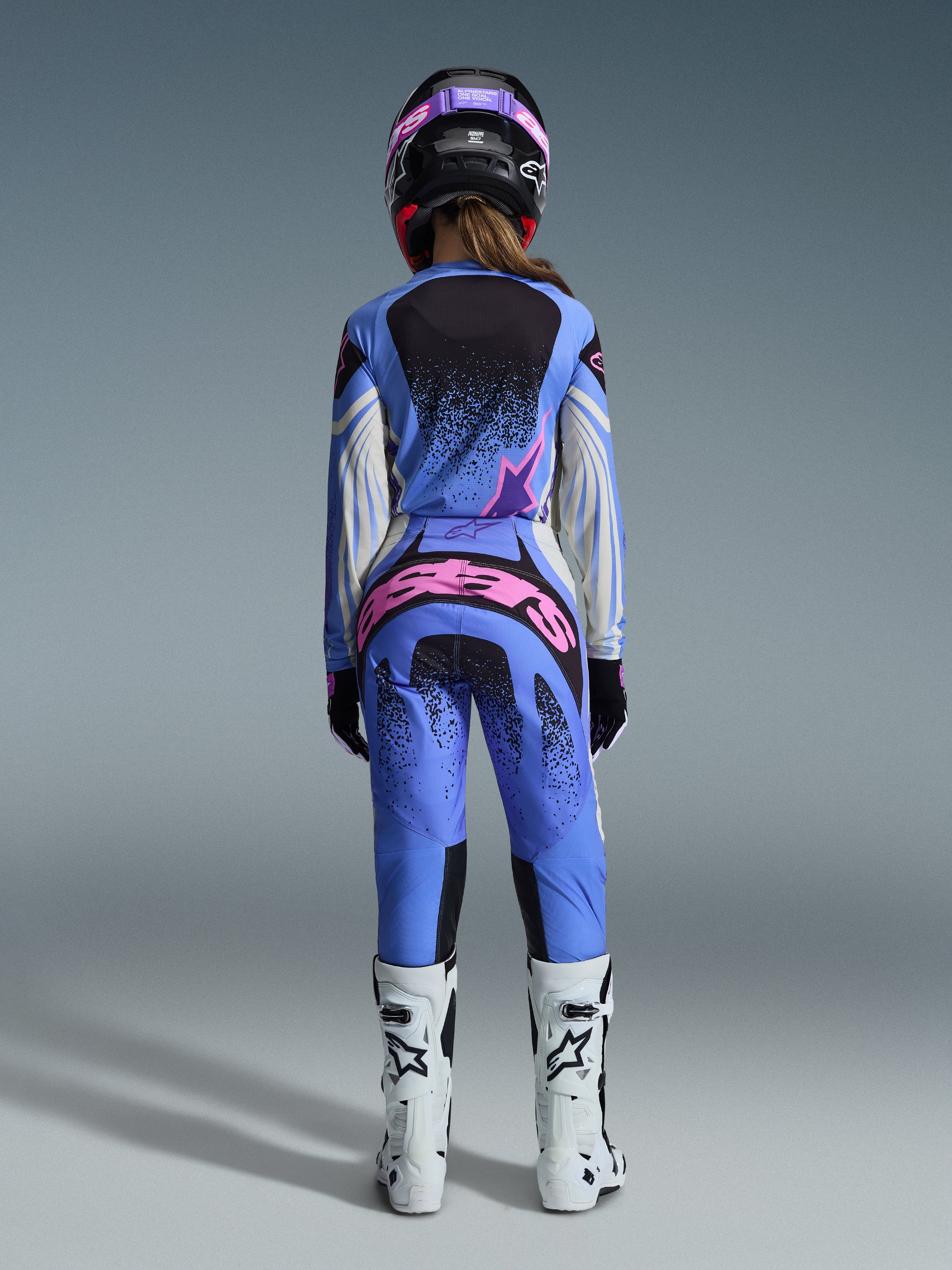 Alpinestars 2026 Stella Techstar Nomur-udstyrssæt, set bagfra af motocrosskører, blå trøje og bukser med lyserøde og hvide detaljer, markant lyserød branding bag på bukserne, hvide motocross-støvler og koordineret sort og lyserød hjelm, professionelt motocross-udstyr