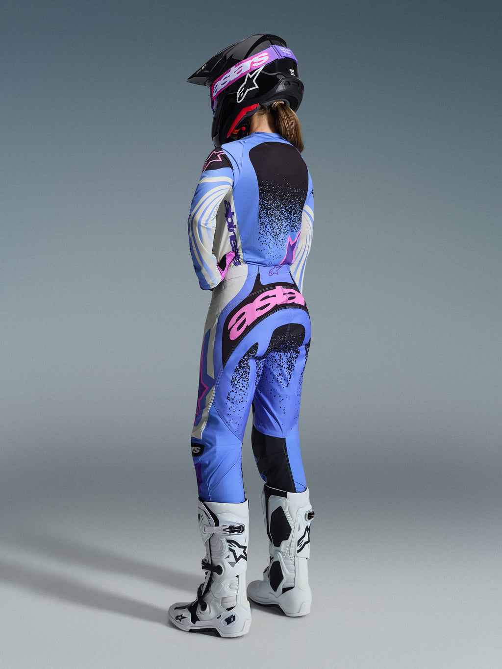 Alpinestars 2026 Stella Techstar Nomur Gear Set, set bagfra af en kvindelig motocrosskører iført et lyseblåt, hvidt og lyserødt jersey- og buksesæt med sorte plettede detaljer, kombineret med en sort hjelm og hvide støvler, professionelt racerudstyr til kvinder.