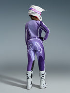 Alpinestars 2026 Stella Fluid Apex Gear Set, motocross-udstyr til kvinder, lilla og lavendel med hvide detaljer, kvindelig kører set bagfra iført matchende trøje og bukser, hvid hjelm med pink goggles og hvide støvler, koordineret atletisk raceroutfit