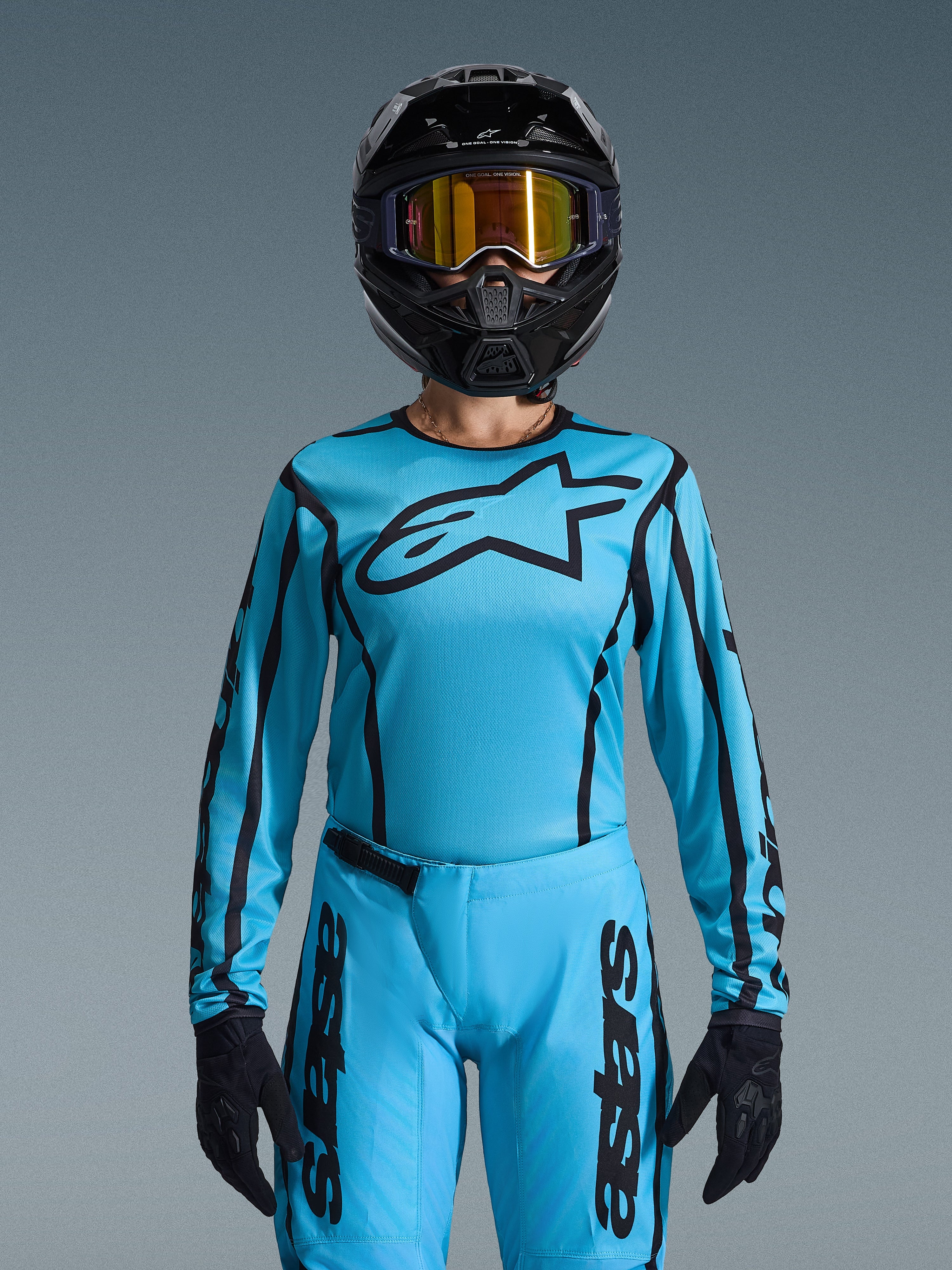 Motocross-kører iført Alpinestars 2026 Stella Fluid Apex-udstyrssæt til damer, lyseblå trøje med sorte konturer og logo, matchende bukser, sort hjelm med iriserende guld-goggles og sorte handsker, koordineret motocross-raceroutfit til kvinder