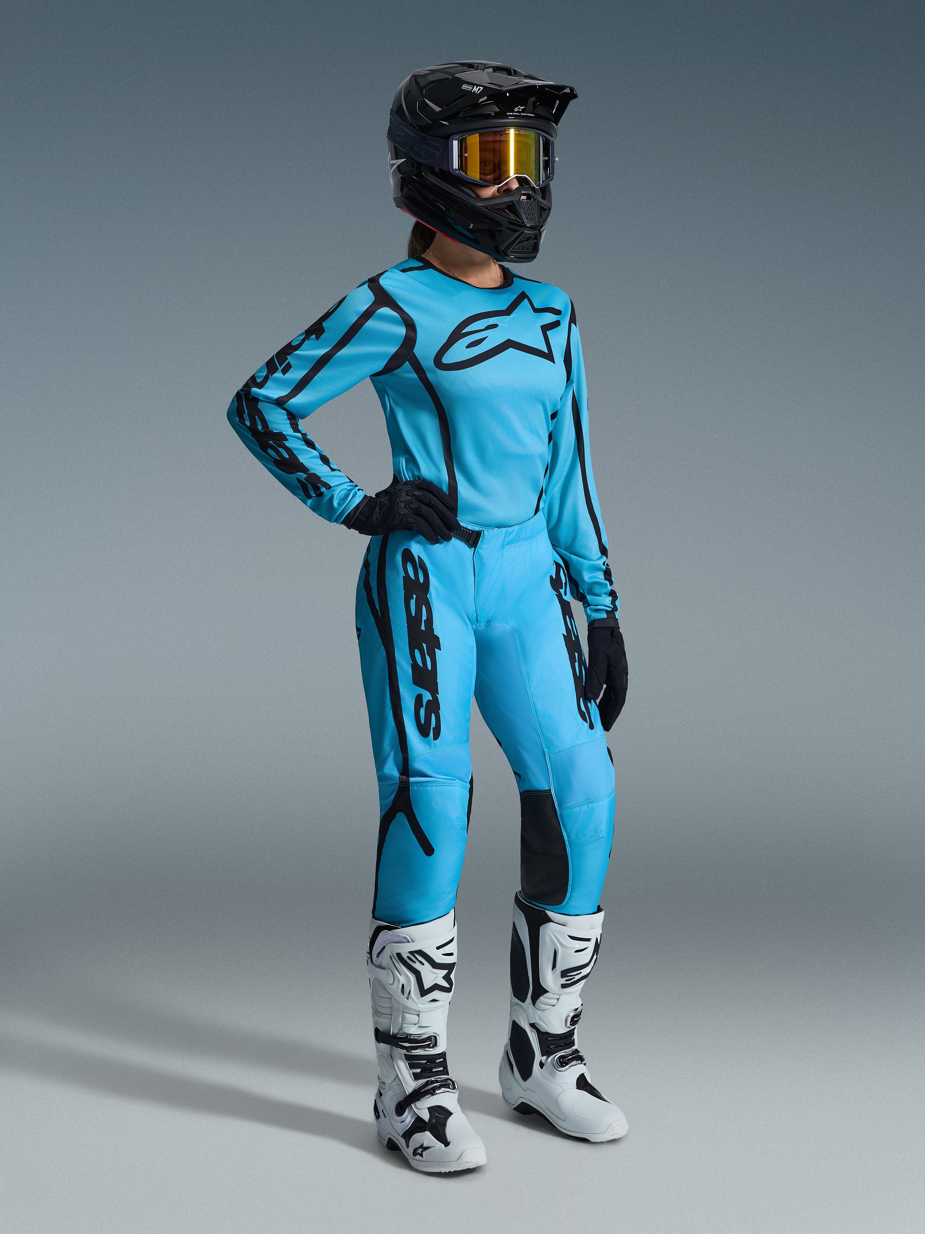 Alpinestars 2026 Stella Fluid Apex-udstyrssæt til damer i farven Tropical Black, med en lyseblå trøje og matchende bukser med sorte detaljer, båret af en kvindelig kører med sort hjelm, iriserende goggles og hvide støvler, professionelt motocross-racerudstyr.