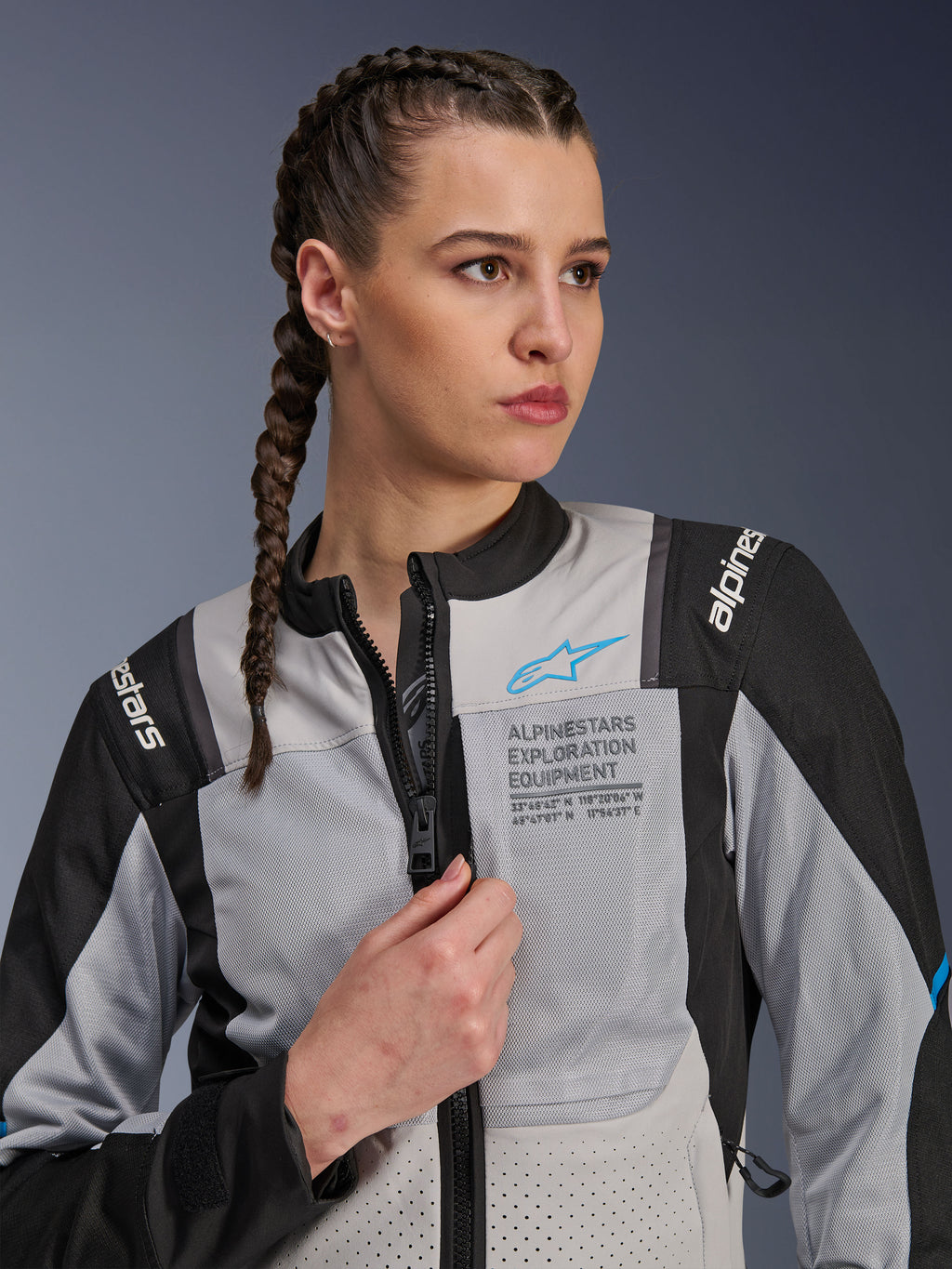 Kvinde iført Alpinestars Stella ST-2 Air-jakke i Ice Gray og sort, nærbillede-portræt, med mesh-paneler til ventilation, 'ALPINESTARS EXPLORATION EQUIPMENT'-branding på brystet med et blåt logo, moderne adventure-motorcykeljakke