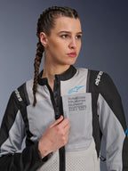 Kvinde iført Alpinestars Stella ST-2 Air-jakke i Ice Gray og sort, nærbillede-portræt, med mesh-paneler til ventilation, 'ALPINESTARS EXPLORATION EQUIPMENT'-branding på brystet med et blåt logo, moderne adventure-motorcykeljakke