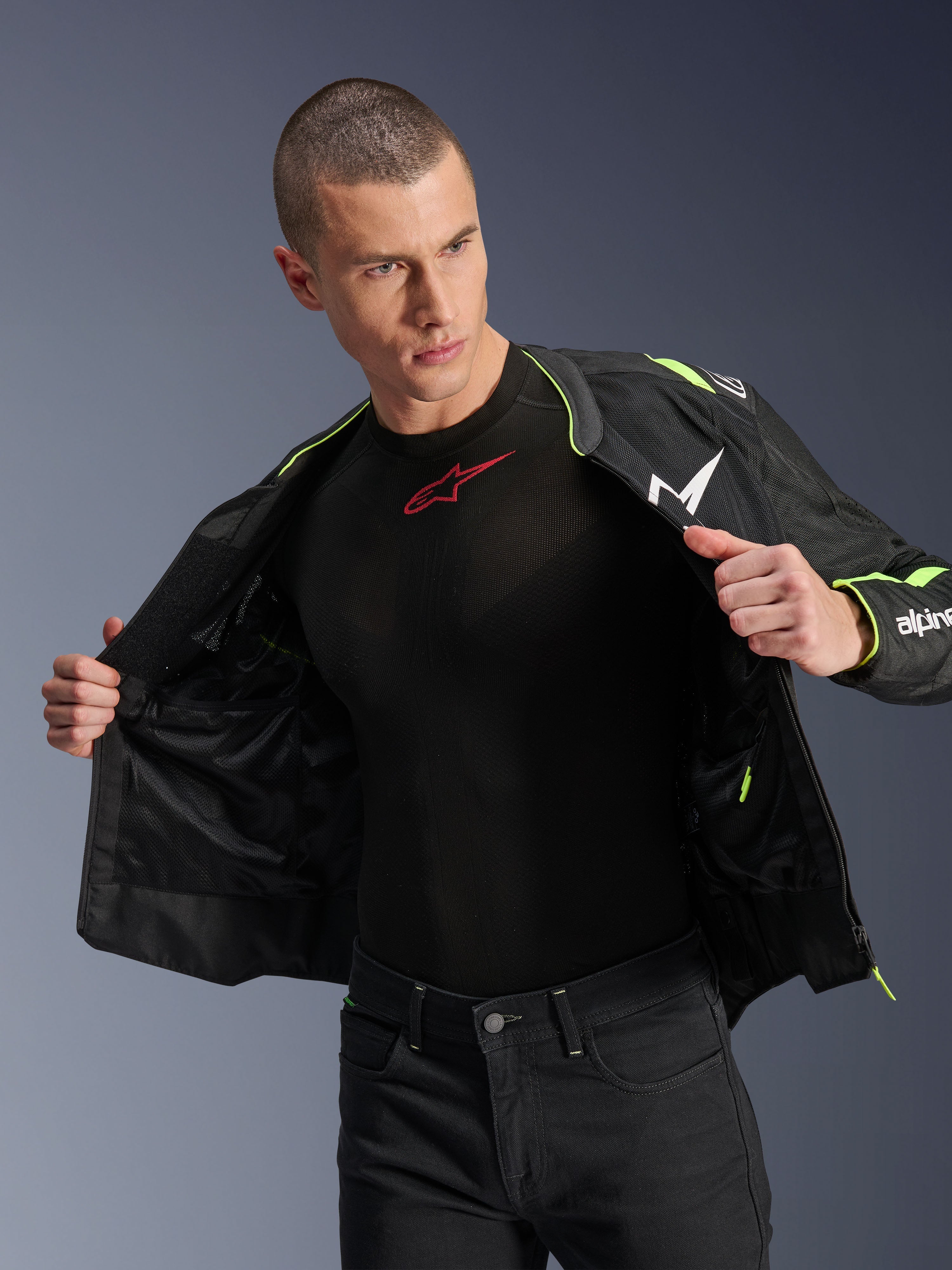 Mandlig model iført Alpinestars T-Sps Air v2-jakke i sort og gul fluo, vist åben for at fremhæve den indvendige mesh-konstruktion og lommer, kombineret med et sort Alpinestars baselayer og sorte bukser, urban motorcykeludstyr-stil