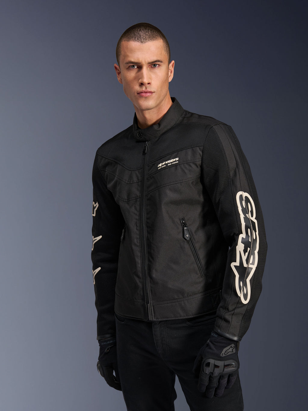 Alpinestars T-Dyno Air-jakke i sort, mandlig model iført en vintage-inspireret tekstil-motorcykeljakke med mesh-paneler til ventilation, stort hvidt Alpinestars-logo på ærmerne, kombineret med sorte kørehandsker, urbant motorcykeludstyrsdesign