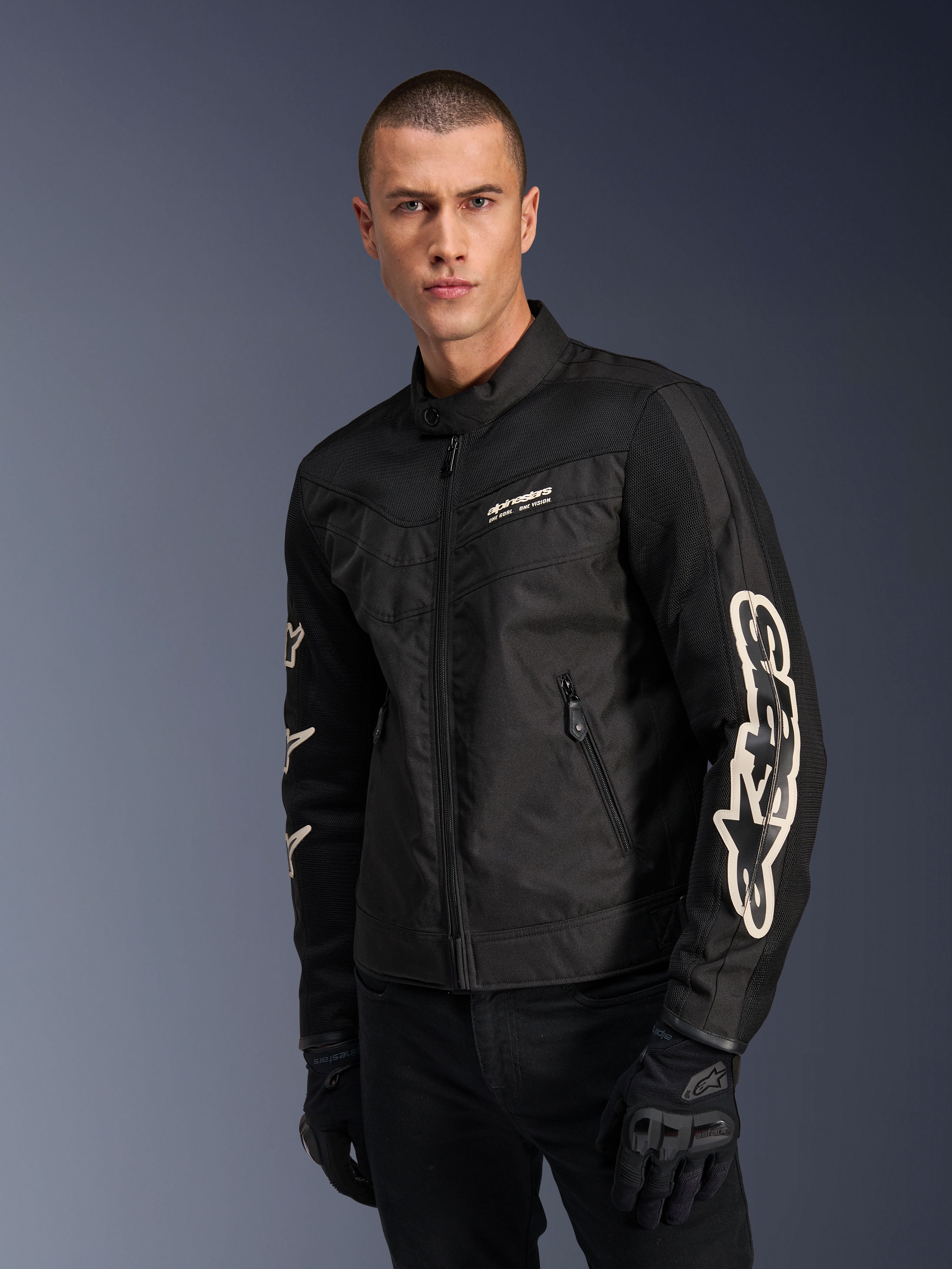 Alpinestars T-Dyno Air-jakke i sort, mandlig model iført en vintage-inspireret tekstil-motorcykeljakke med mesh-paneler til ventilation, stort hvidt Alpinestars-logo på ærmerne, kombineret med sorte kørehandsker, urbant motorcykeludstyrsdesign