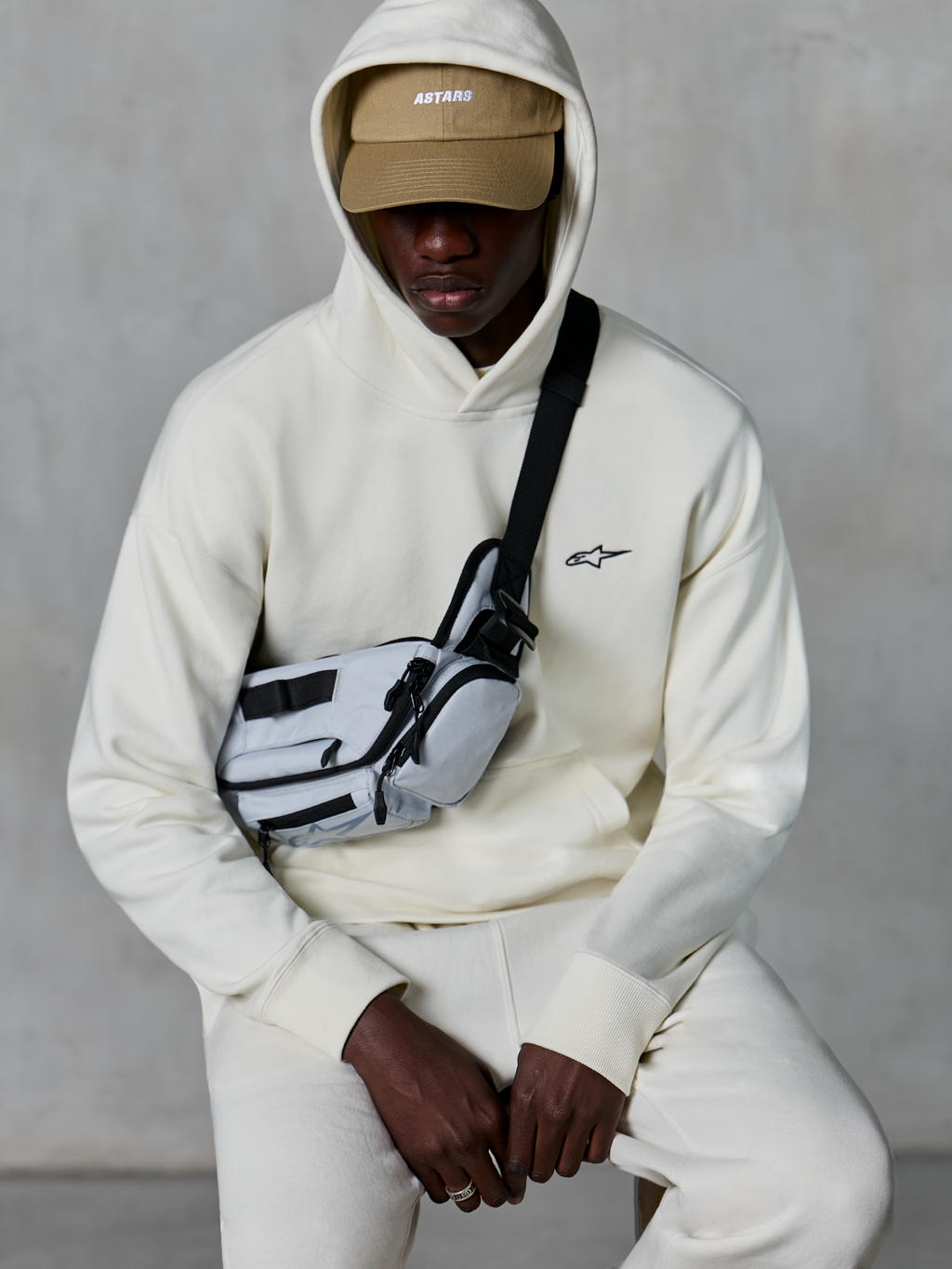 Alpinestars Essentials Ageless hættetrøje, afslappet streetwear, Off White, lille sort broderet logo på brystet, hætte båret over en tan-farvet kasket med ASTARS-branding, stylet med en lysegrå cross-body taske, eksklusivt lifestyle-tøj