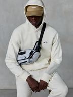 Alpinestars Essentials Ageless hættetrøje, afslappet streetwear, Off White, lille sort broderet logo på brystet, hætte båret over en tan-farvet kasket med ASTARS-branding, stylet med en lysegrå cross-body taske, eksklusivt lifestyle-tøj