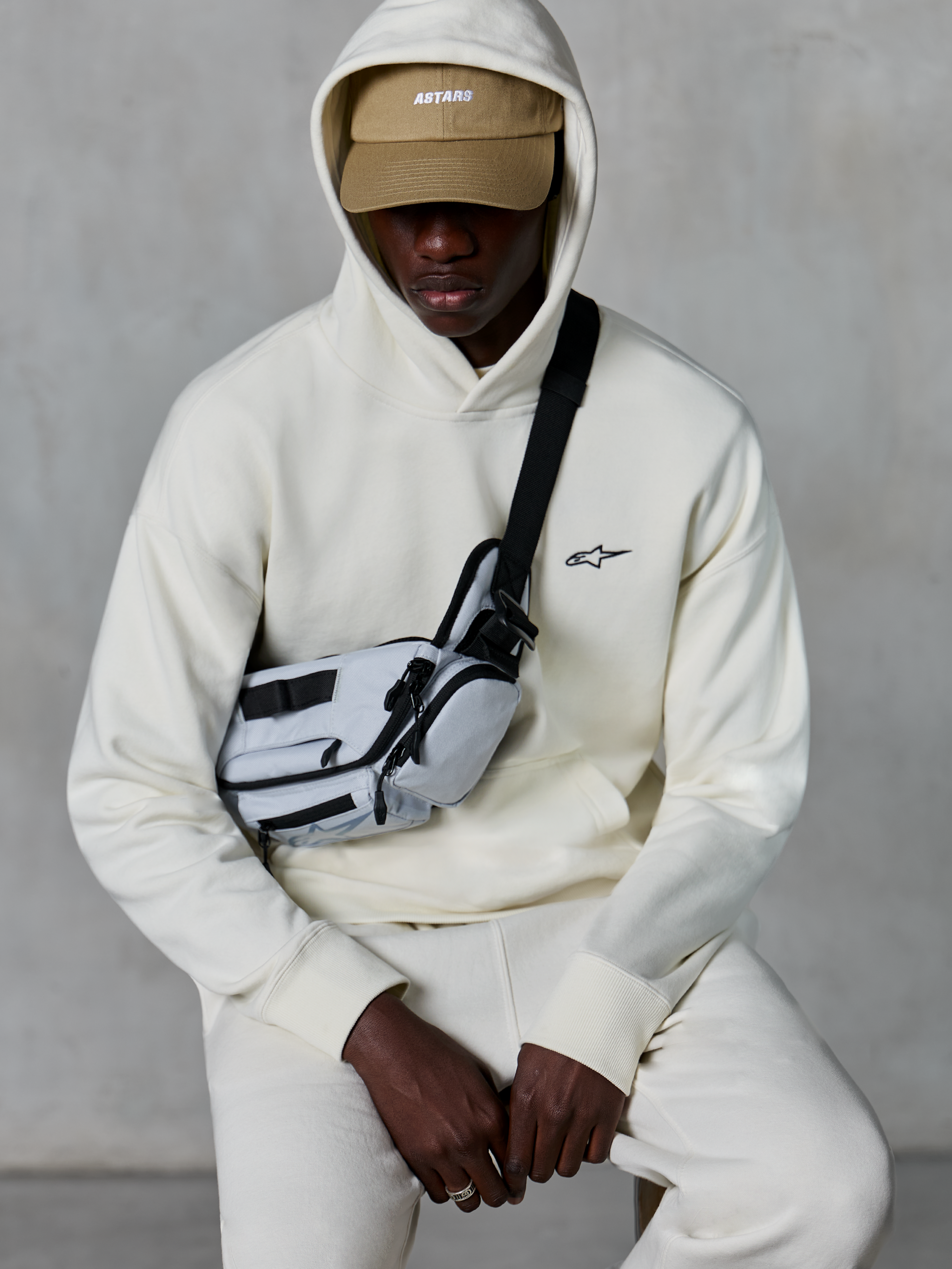 Alpinestars Essentials Ageless hættetrøje, afslappet streetwear, Off White, lille sort broderet logo på brystet, hætte båret over en tan-farvet kasket med ASTARS-branding, stylet med en lysegrå cross-body taske, eksklusivt lifestyle-tøj