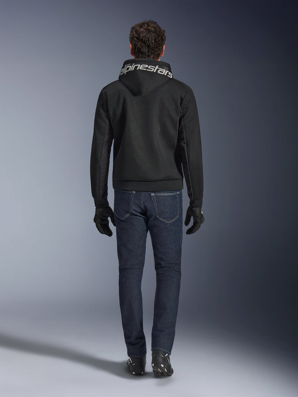 Alpinestars Chrome Superairflow Hoodie, sort motorcykelhoodie med hvid branding på hætten, set bagfra, båret med mørkeblå jeans, sorte handsker og sorte og hvide støvler, urbant sportskøreudstyr