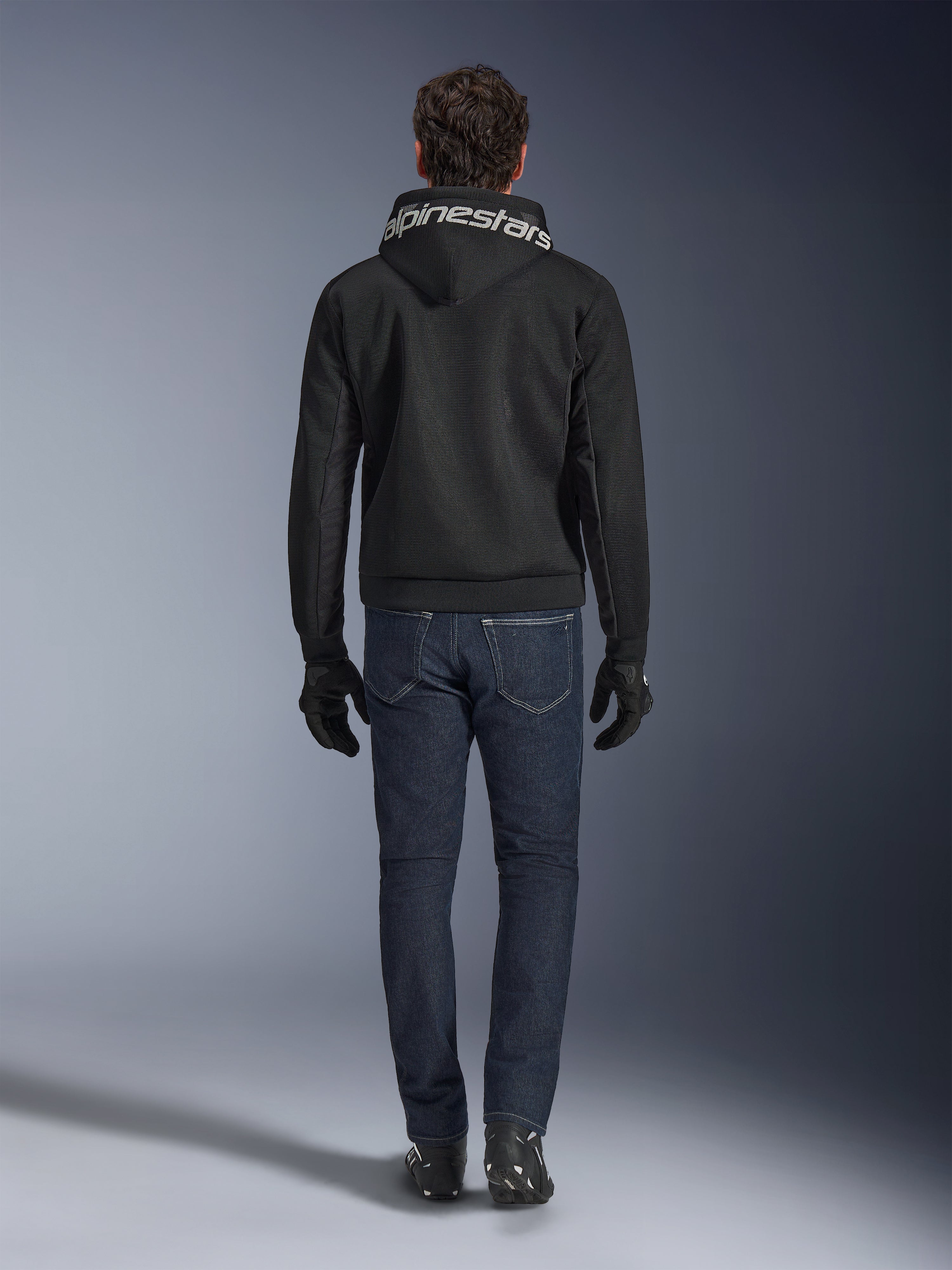 Alpinestars Chrome Superairflow Hoodie, sort motorcykelhoodie med hvid branding på hætten, set bagfra, båret med mørkeblå jeans, sorte handsker og sorte og hvide støvler, urbant sportskøreudstyr