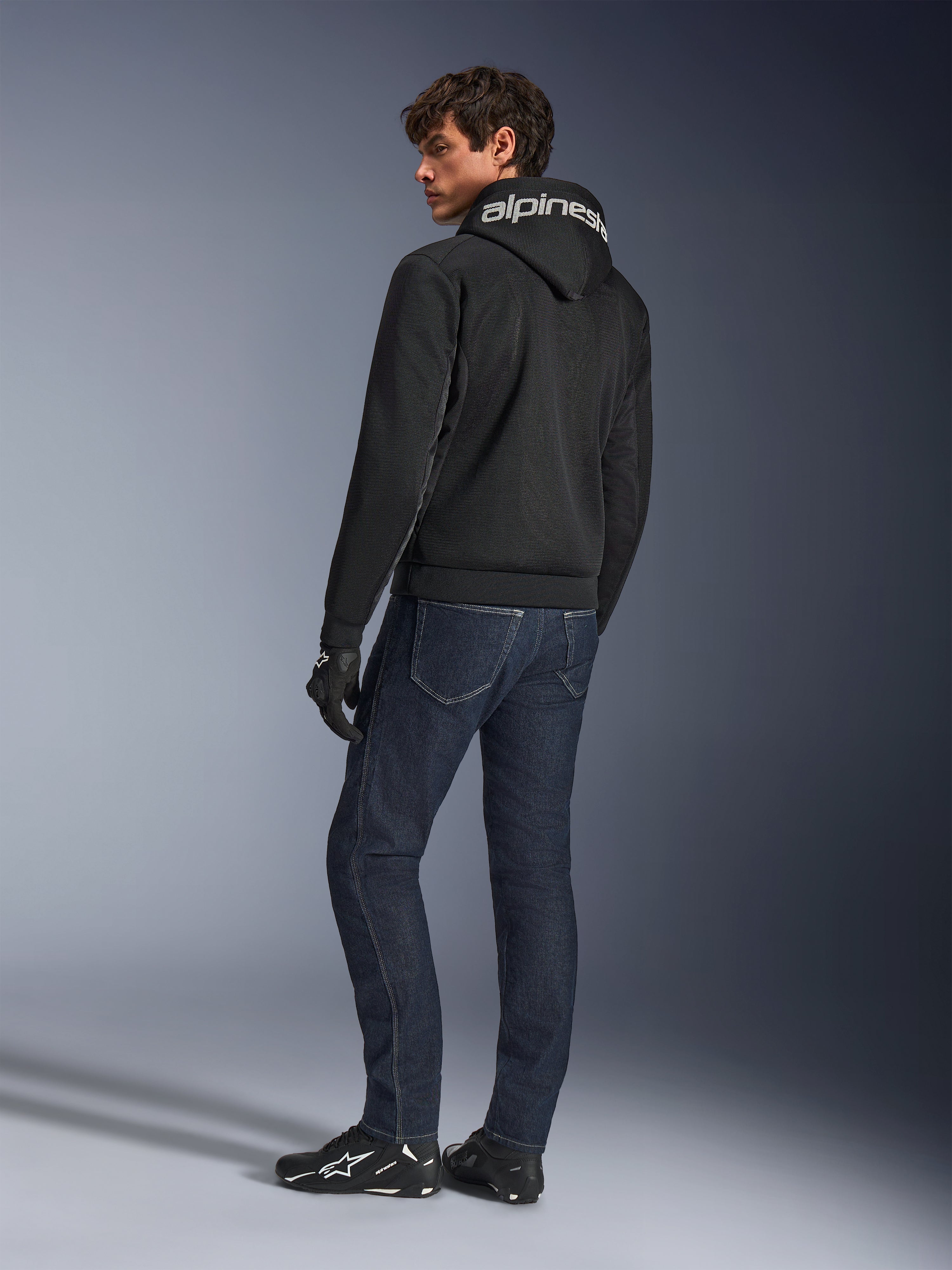 Alpinestars Chrome Superairflow Hoodie, sort, set bagfra på mandlig model, med hvidt Alpinestars-logo på hætten, båret med mørke denimjeans og sorte handsker, urbant motorcykeludstyr