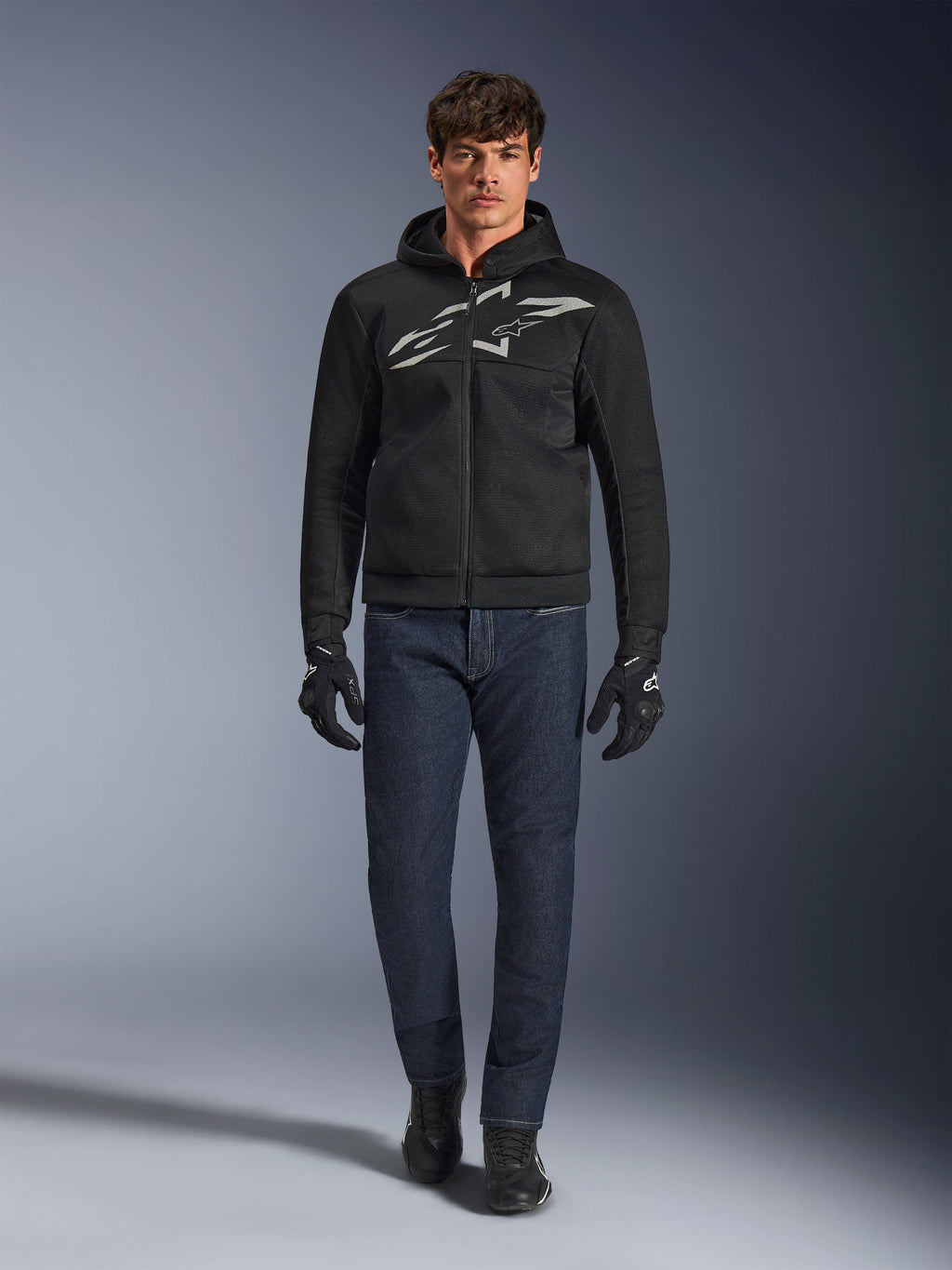 Motorcykelrytter iført Alpinestars Chrome Superairflow Hoodie i sort og grå, kombineret med mørkeblå jeans, sorte motorcykelhandsker og sorte sko. Hættetrøjen har et stort stiliseret logo på brystet og en 3D-mesh-tekstur til ydeevne ved høje temperaturer.