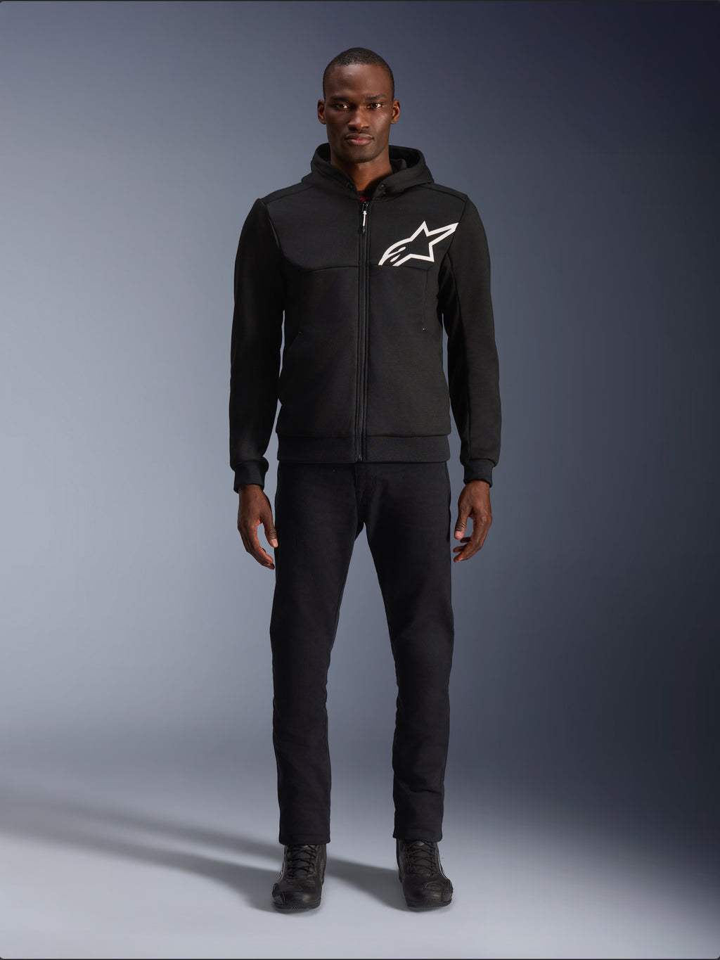 Alpinestars Chrome V2 Sport Hoodie, sort og hvid motorcykel-hættetrøje, med et stort hvidt Alpinestars-logo på brystet, lynlås foran og hætte, båret af en model i sorte bukser og støvler, beskyttende køreudstyr i urban stil.