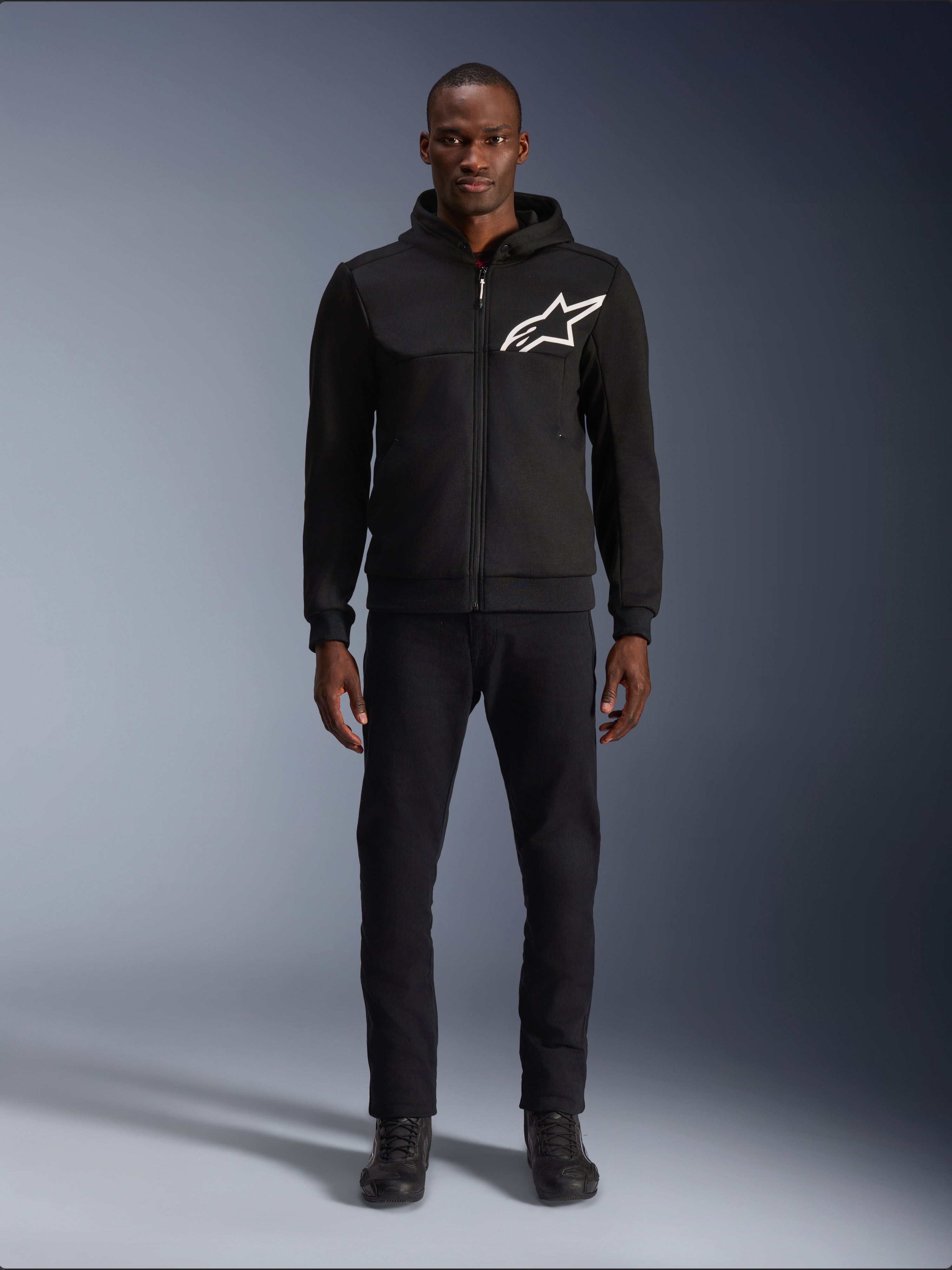 Alpinestars Chrome V2 Sport Hoodie, sort og hvid motorcykel-hættetrøje, med et stort hvidt Alpinestars-logo på brystet, lynlås foran og hætte, båret af en model i sorte bukser og støvler, beskyttende køreudstyr i urban stil.