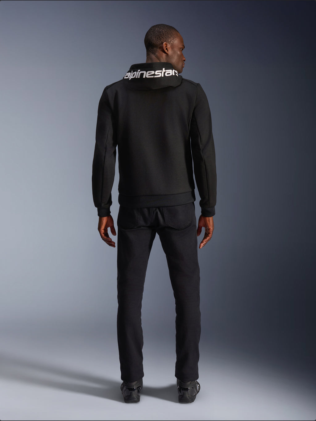 Alpinestars Chrome V2 Sport Hoodie, motorcykel-hættetrøje, sort, model set bagfra iført sort tech fleece-hættetrøje med hvidt Alpinestars-logo på hætten, kombineret med sorte bukser og støvler, afslappet urban kørestil
