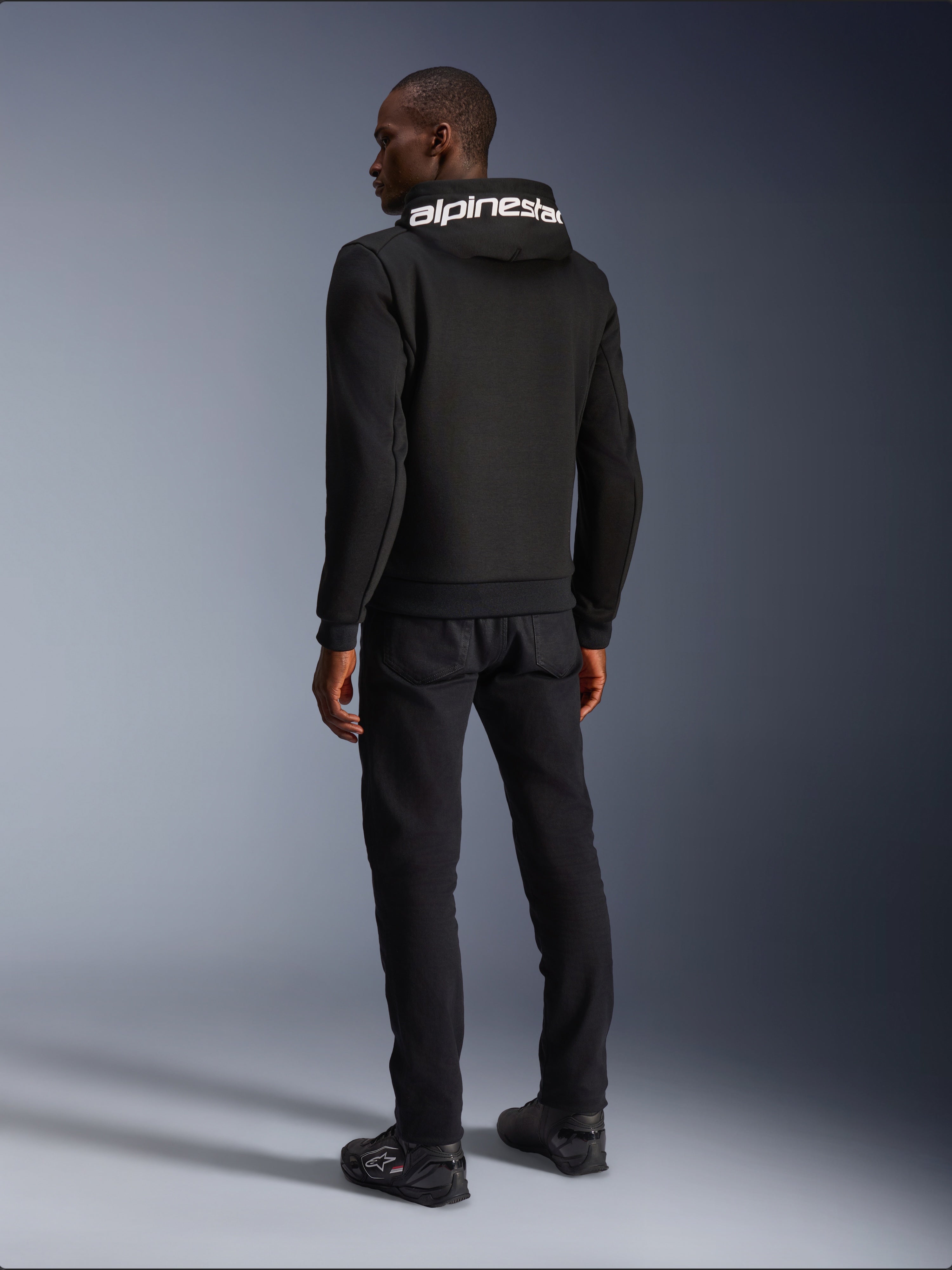 Alpinestars Chrome V2 Sport Hoodie, sort, set bagfra med hvidt Alpinestars-branding på hætten, båret med sorte bukser og køresko, afslappet urban motorcykelbeklædning med en atletisk silhuet