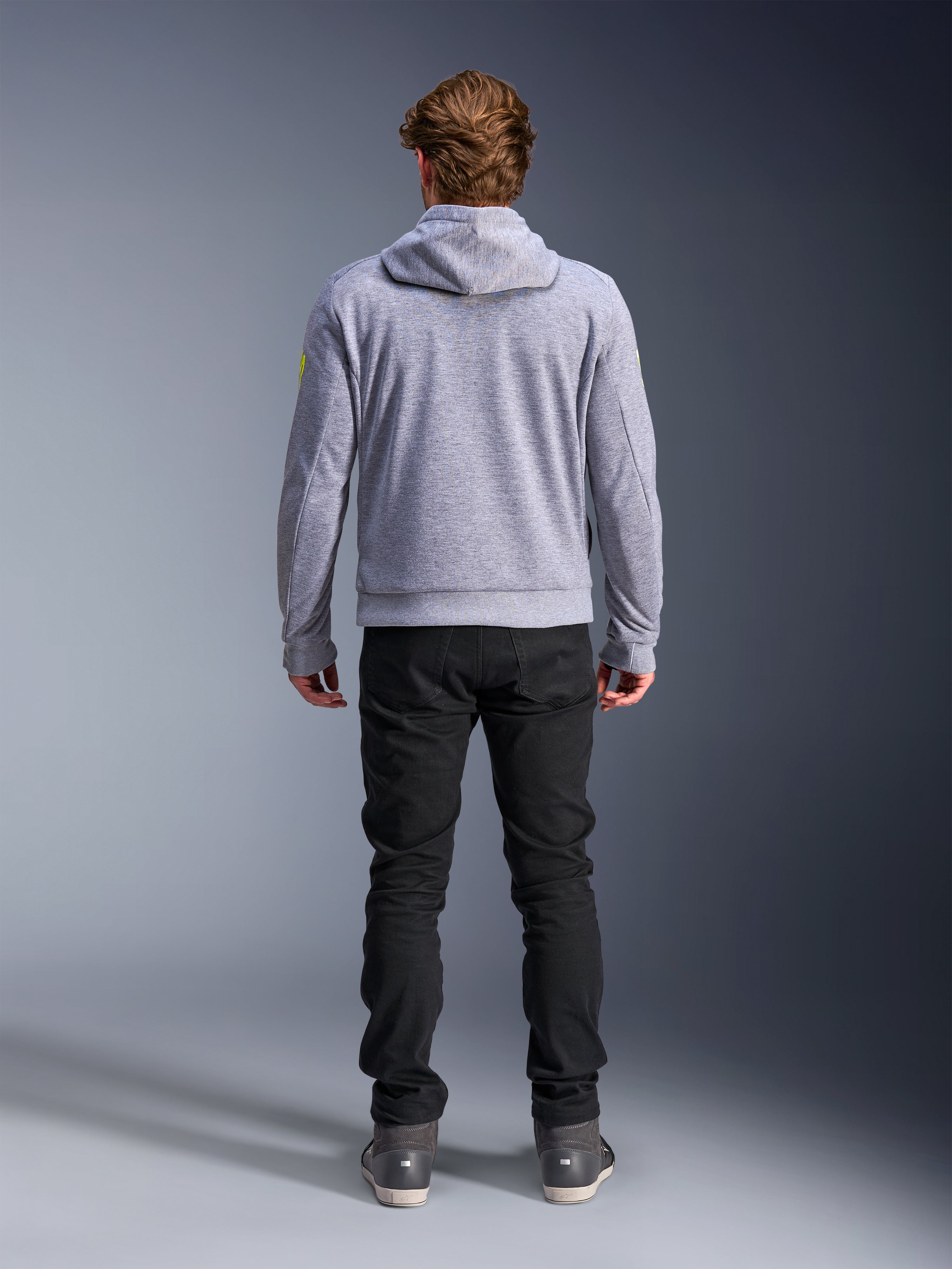 Alpinestars Chrome V2 Sport Hoodie, Gray Heather Fluo Yellow, set bagfra af urbant motorcykeludstyr, tech fleece-hættetrøje båret med sorte bukser, med fluorgule detaljer på ærmerne.