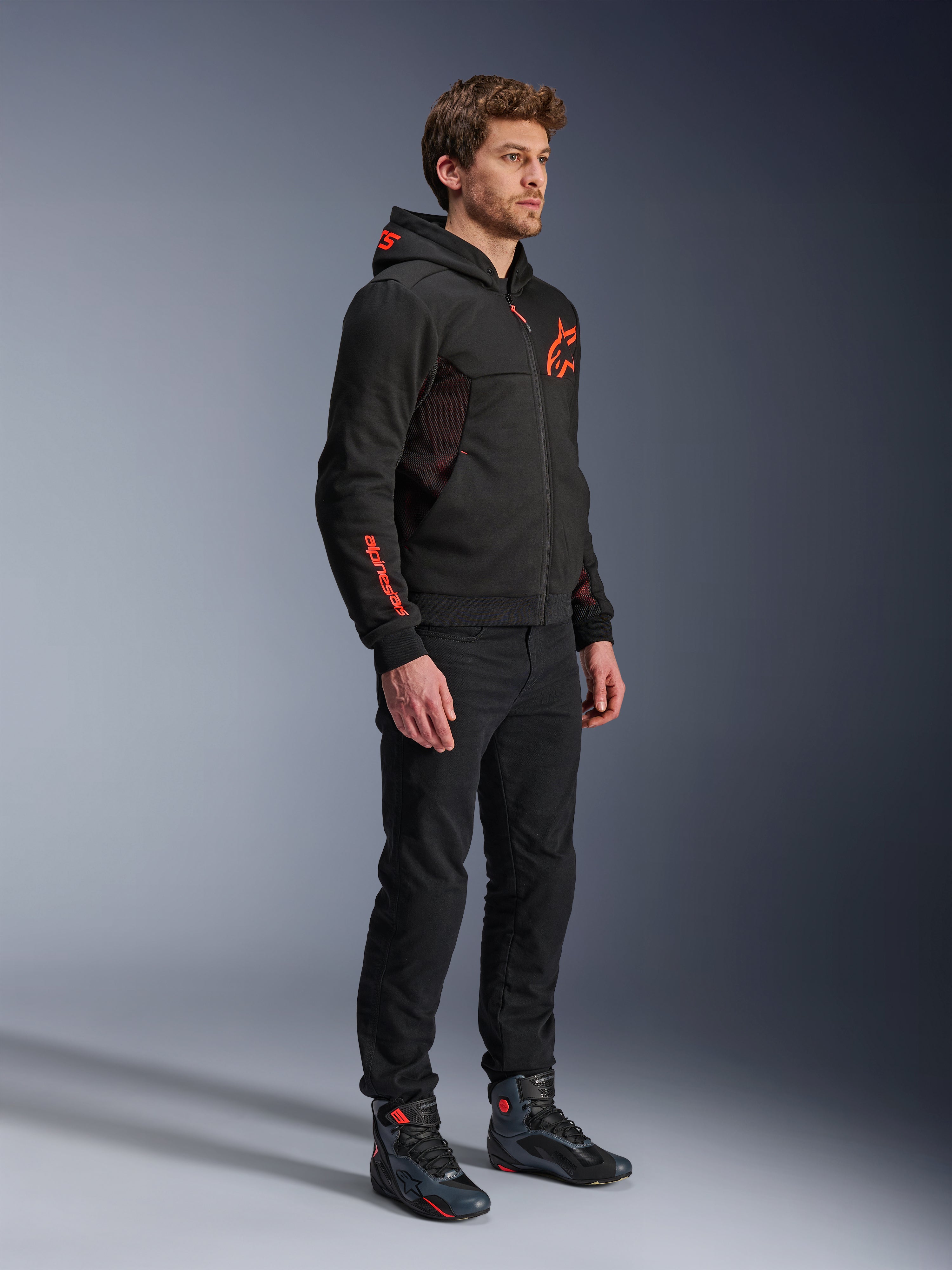 Alpinestars Chrome Air Sport Hoodie, Black Red Fluo, urban motorcykeljakke med tech-fleece, båret med sorte bukser og motorcykelstøvler, med mesh-ventilationspaneler og røde fluorescerende logoer på brystet og ærmet.
