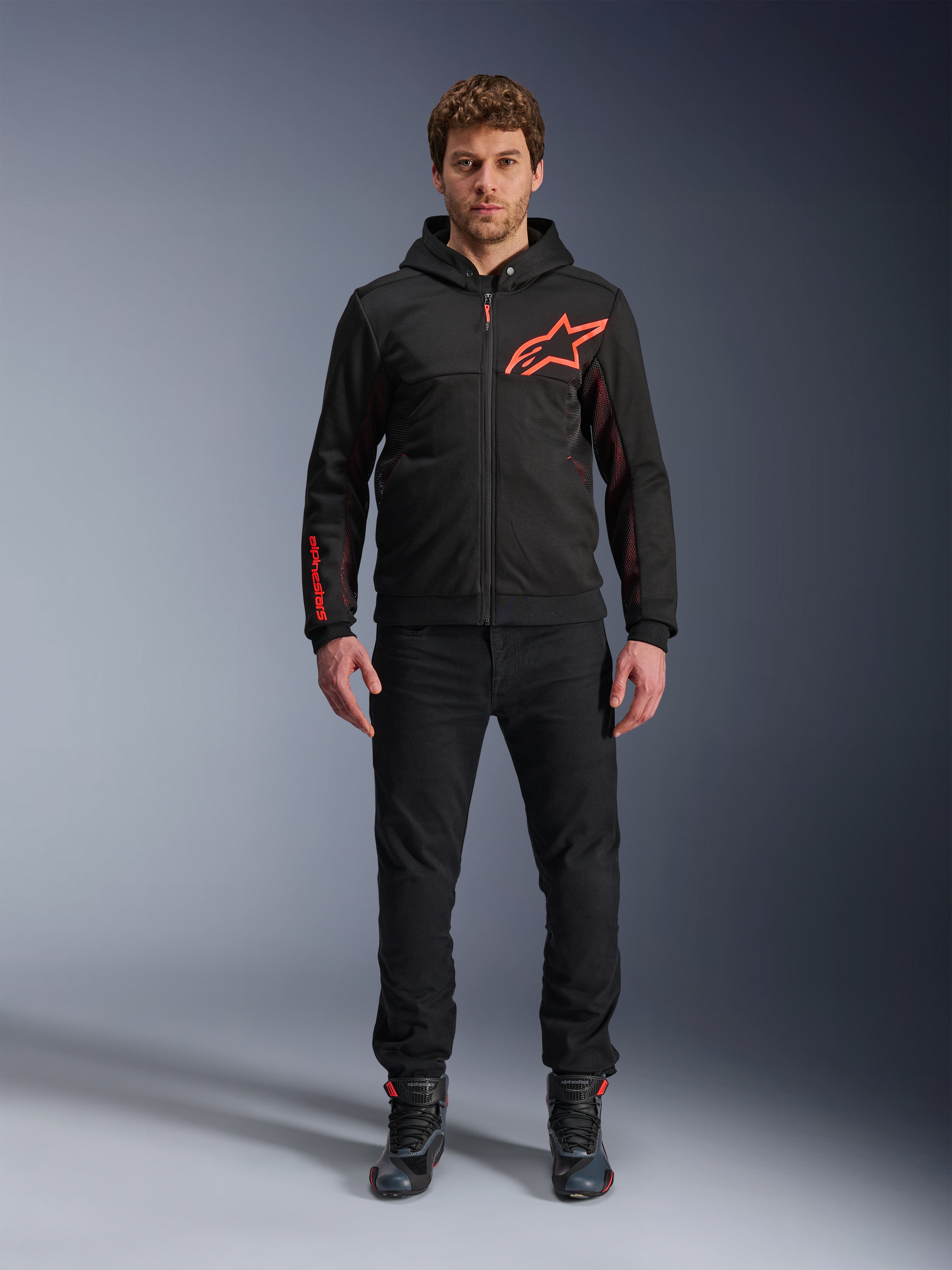 Alpinestars Chrome Air Sport Hoodie, sort og fluorescerende rød, urban motorcykeljakke med tech-fleece-konstruktion, med et stort rødt Astars-logo på brystet og branding på ærmet, båret af en model med sorte bukser og køresko