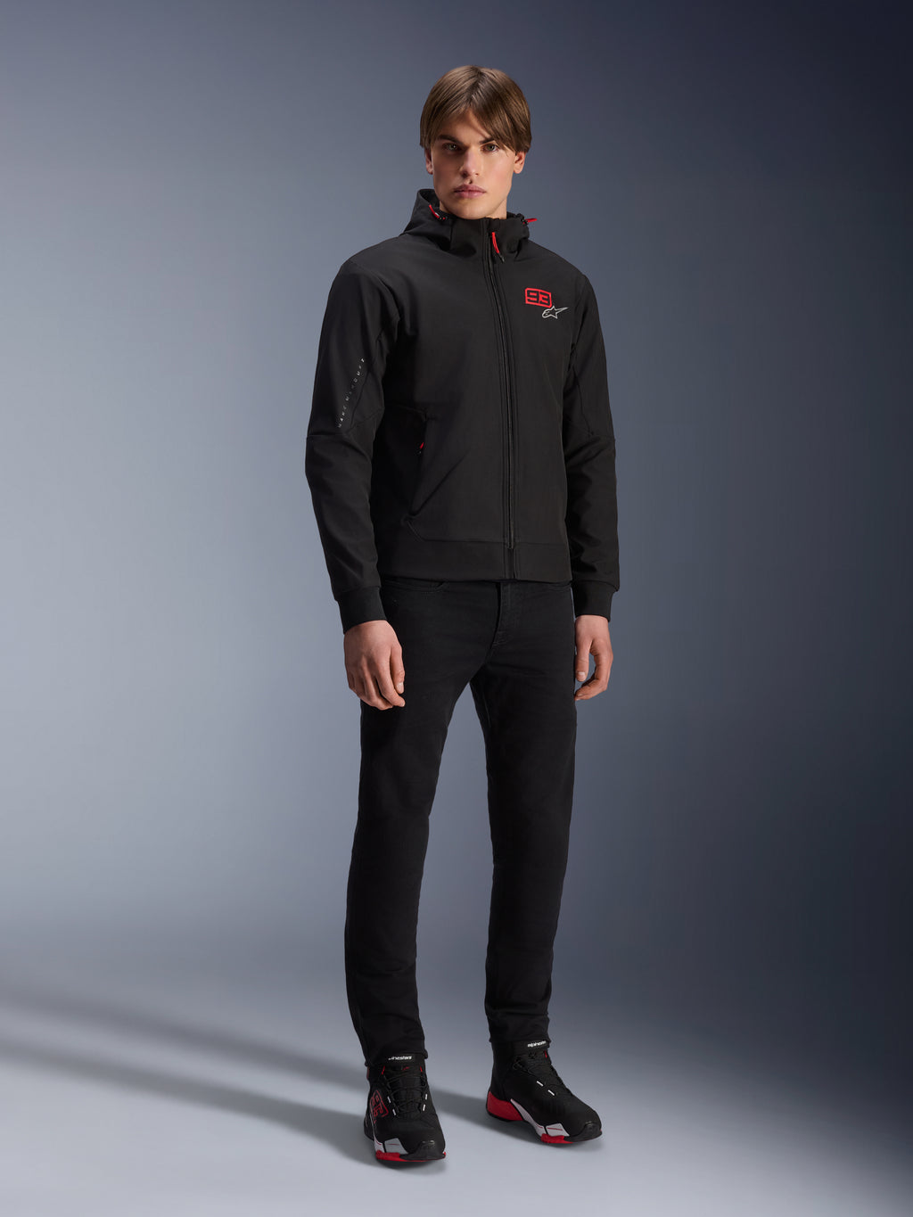 Alpinestars MM93 Radium Tech Hoodie, Urban Motorcycle Jacket, Black with Bright Red accents, worn by model with black pants and riding shoes, featuring red '93' chest logo and Marc Marquez branding on the sleeve, casual technical streetwear for urban riding