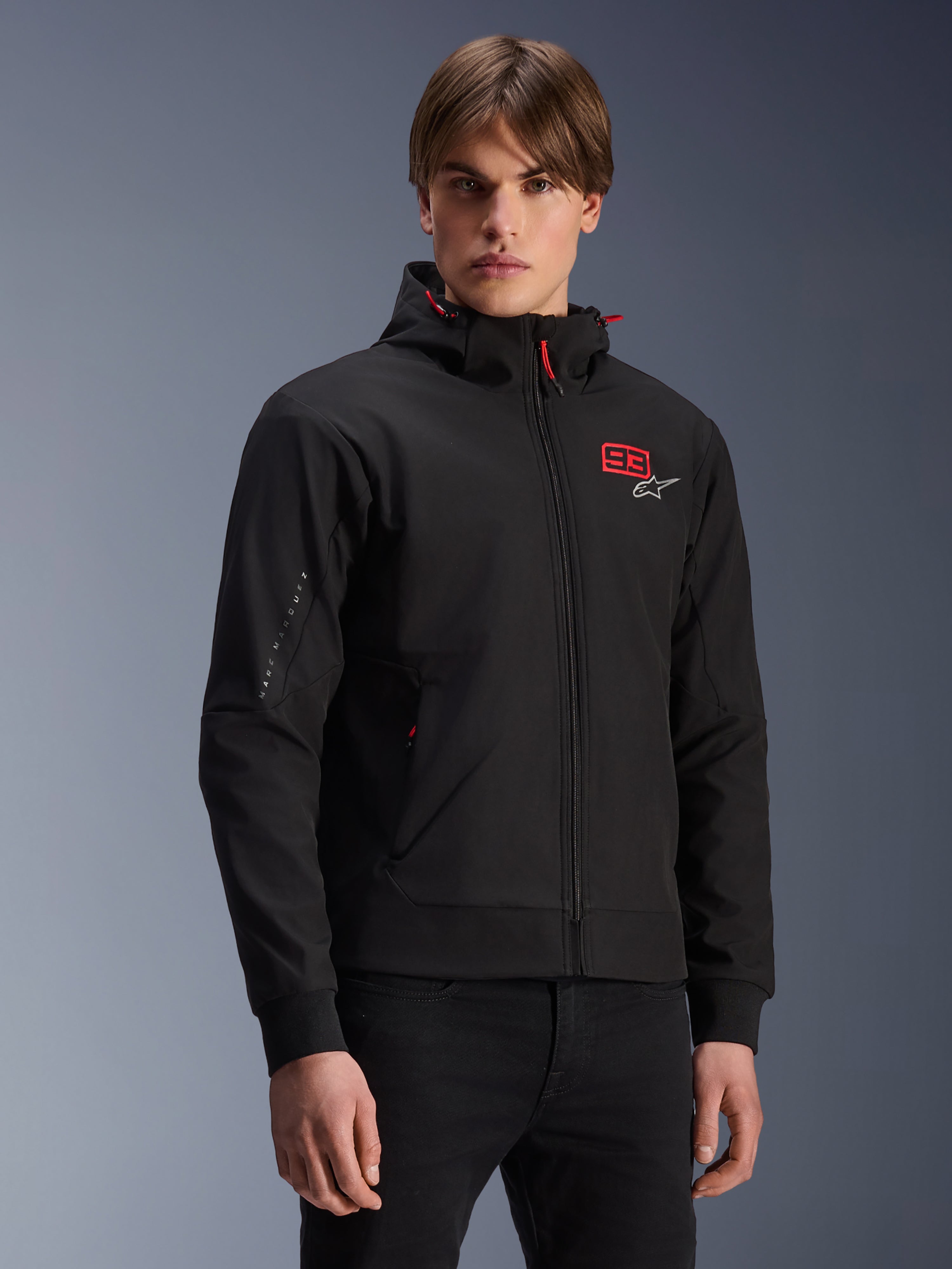 MM93 Radium Tech Hoodie, sort urban motorcykeljakke med lynlås, rød 93-grafik og hvidt Alpinestars-logo på brystet, båret af model med sorte bukser, aftagelig hætte med røde snore og branding på ærmet, afslappet motorsport-inspireret design