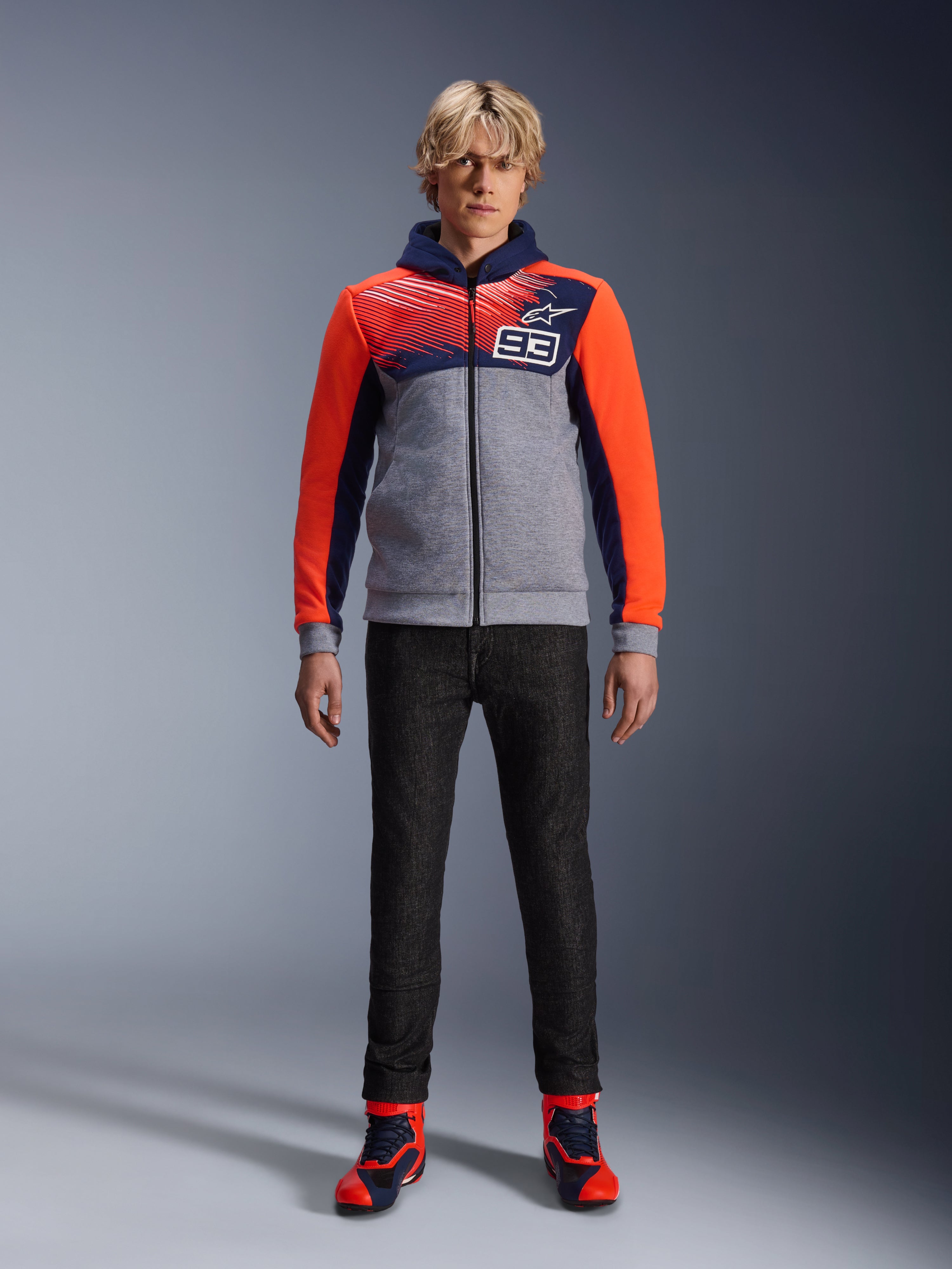 Alpinestars MM93 Chrome V2 Sport Hoodie, motorcykelhættetrøje, Blue Navy, Gray Melange og Red Fluo, båret med mørke kørebukser og sneakers med røde detaljer, med gennemgående lynlås og MM93-logo, afslappet urbant motorsport-inspireret design