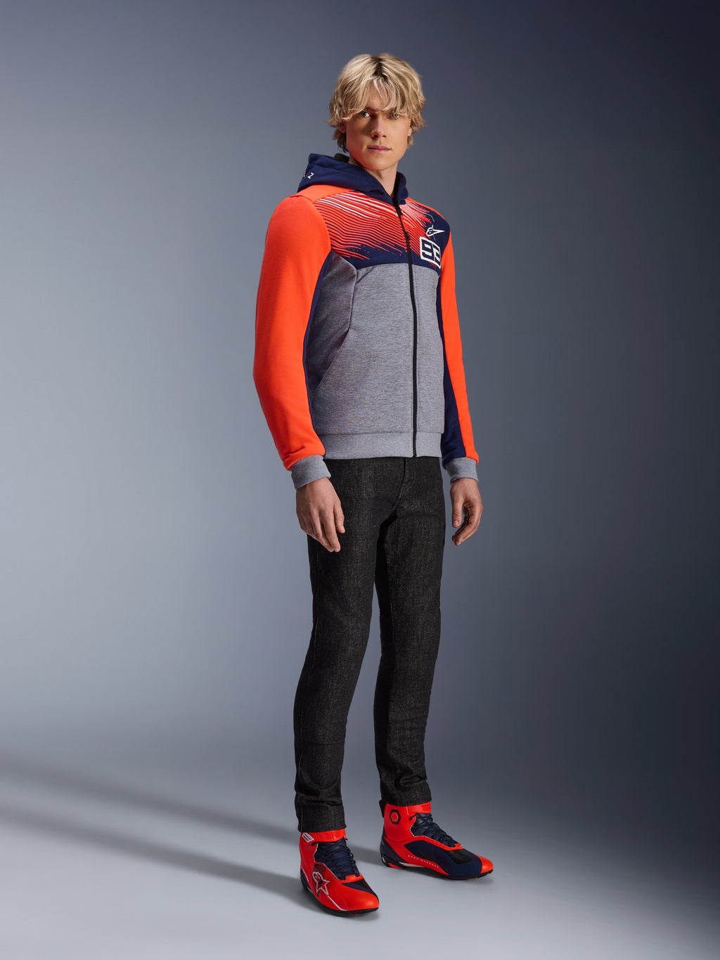 Alpinestars MM93 Chrome V2 Sport Hoodie, urban motorcykel-hættetrøje, gråmeleret krop med navyblåt bryst og neonrøde ærmer, med 93-logoet på brystet, båret af en model med sorte slim-fit bukser og matchende røde og blå high-top køresko, moderne atletisk pasform til bykørsel
