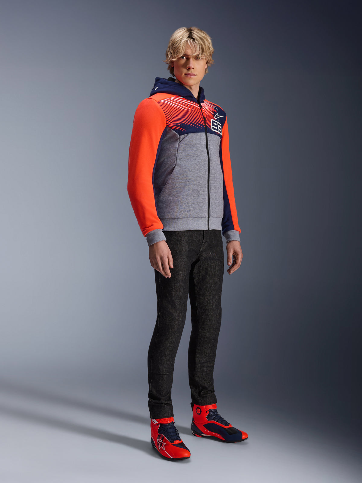 Alpinestars MM93 Chrome V2 Sport Hoodie, urban motorcykel-hættetrøje, gråmeleret krop med navyblåt bryst og neonrøde ærmer, med 93-logoet på brystet, båret af en model med sorte slim-fit bukser og matchende røde og blå high-top køresko, moderne atletisk pasform til bykørsel