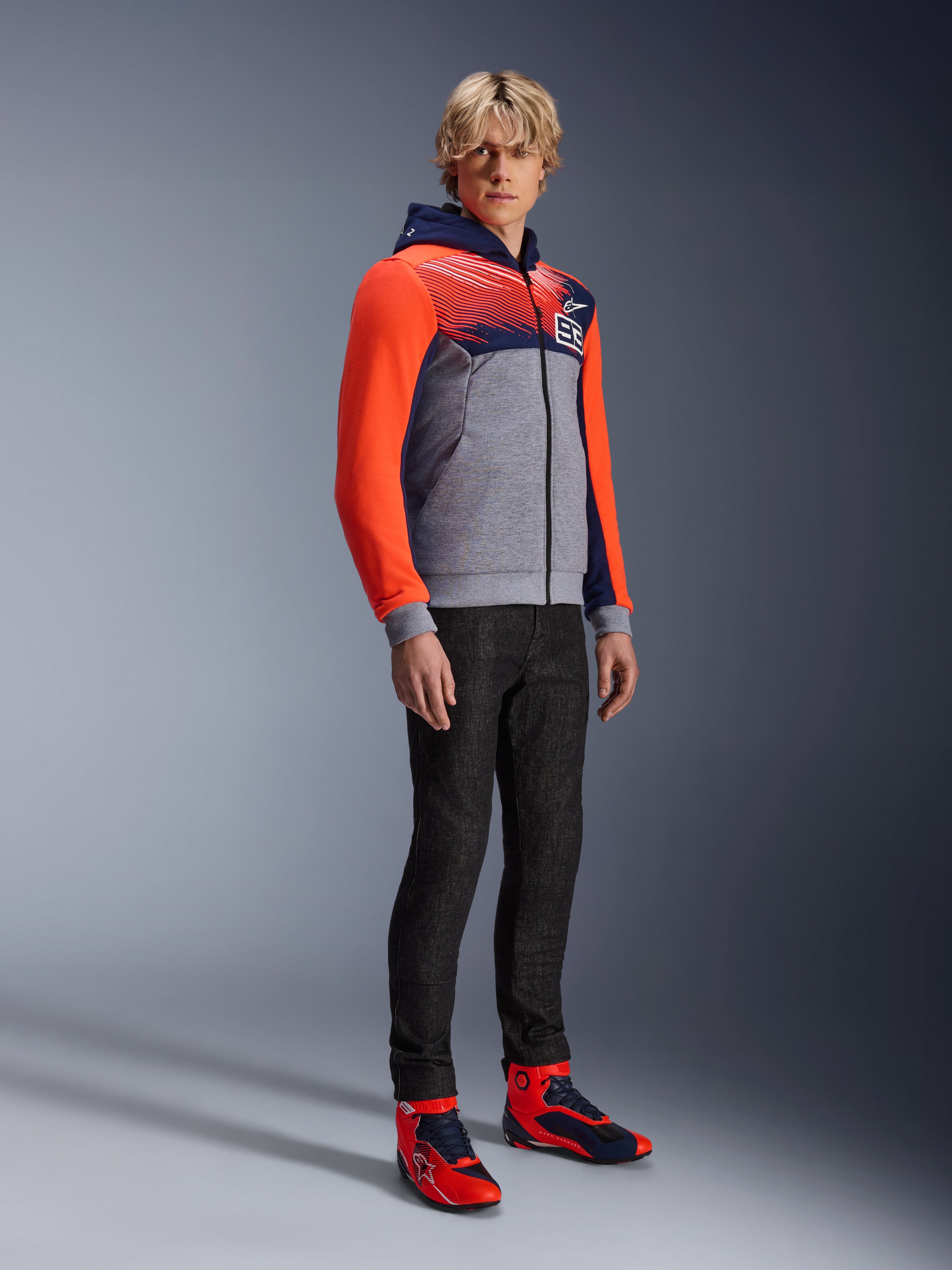 Alpinestars MM93 Chrome V2 Sport Hoodie, urban motorcykel-hættetrøje, gråmeleret krop med navyblåt bryst og neonrøde ærmer, med 93-logoet på brystet, båret af en model med sorte slim-fit bukser og matchende røde og blå high-top køresko, moderne atletisk pasform til bykørsel