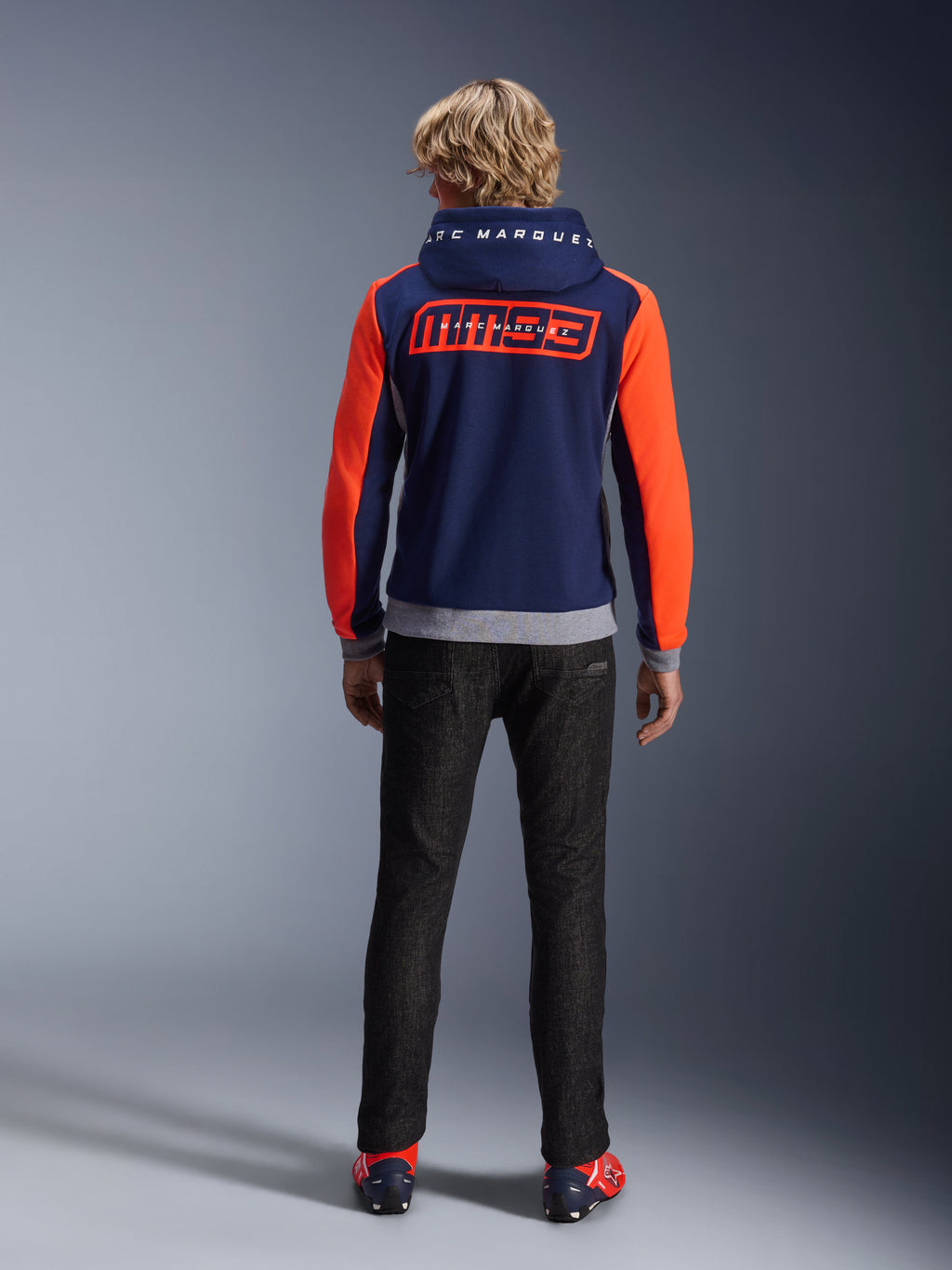 Model set bagfra iført Alpinestars MM93 Chrome V2 Sport Hoodie, mørkeblå med stærke neonrøde ærmer og grå melerede detaljer, store MM93 og Marc Marquez logoer på ryggen og hætten, afslappet motorcykel-hættetrøje kombineret med sorte bukser