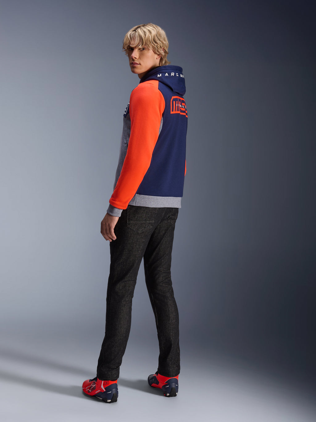 Alpinestars MM93 Chrome V2 Sport Hættetrøje, Blue Navy Gray Mel Red Fluo, model iført hættetrøje med klare røde ærmer og MM93 Marc Marquez-branding på ryggen, stylet med mørke jeans og røde køresko, afslappet urbant motorcykeludstyr