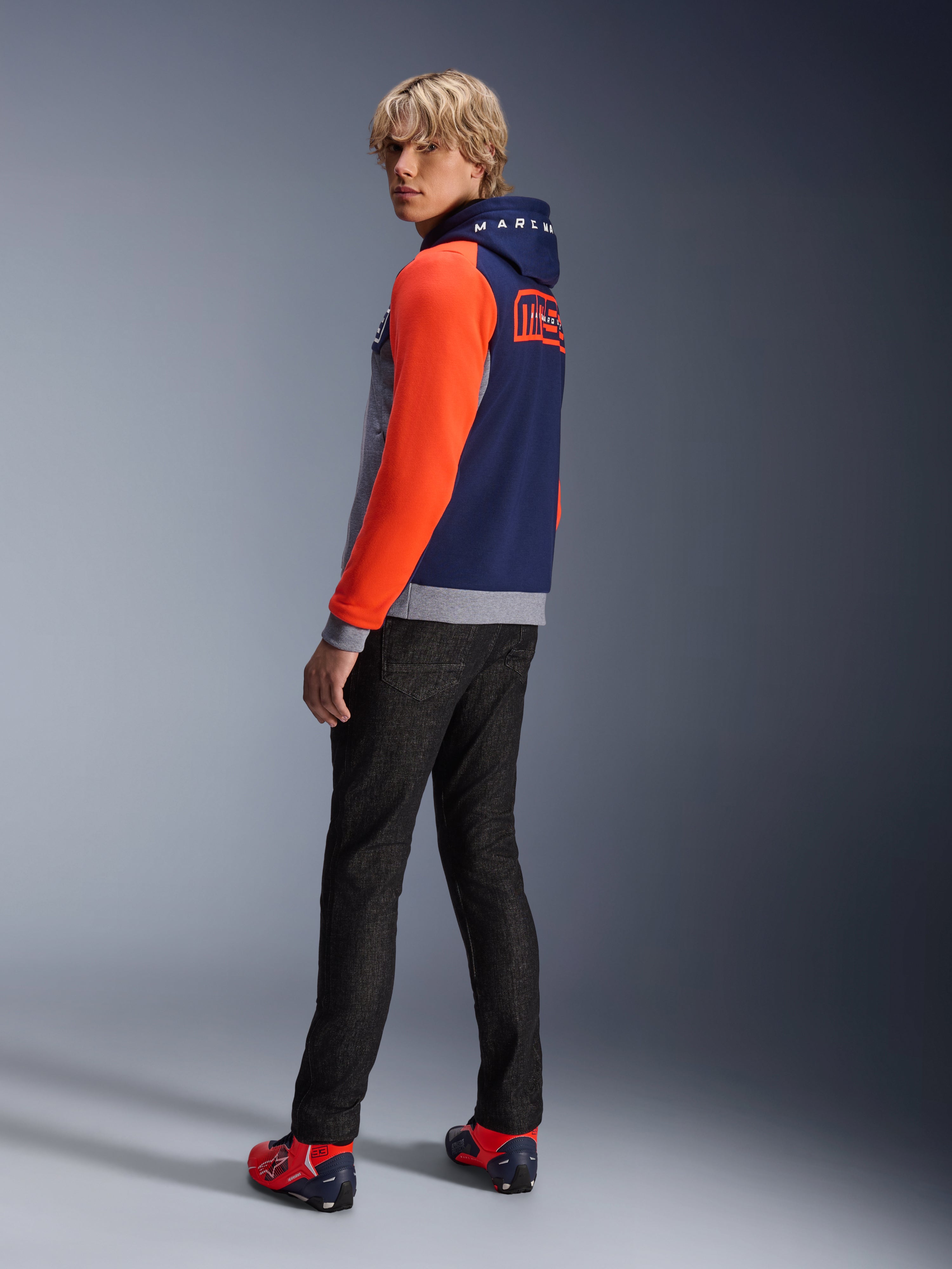 Alpinestars MM93 Chrome V2 Sport Hættetrøje, Blue Navy Gray Mel Red Fluo, model iført hættetrøje med klare røde ærmer og MM93 Marc Marquez-branding på ryggen, stylet med mørke jeans og røde køresko, afslappet urbant motorcykeludstyr