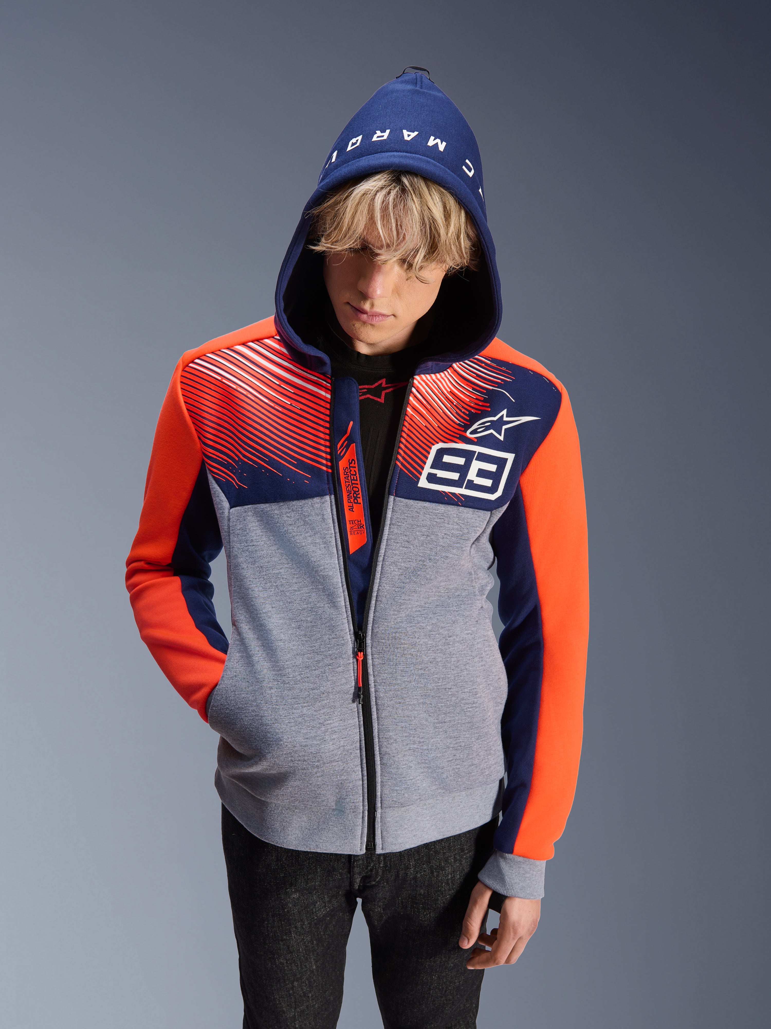 Alpinestars MM93 Chrome V2 Sport hættetrøje, motorcykel hættetrøje, grå meleret krop med mørkeblå overdel og fluorescerende orange ærmer, med hvidt nummer 93 og Alpinestars stjerne-logo, Marc Marquez-tekst på hætten, bæres af en mandlig model med hætten oppe, urbant motorsport-inspireret design