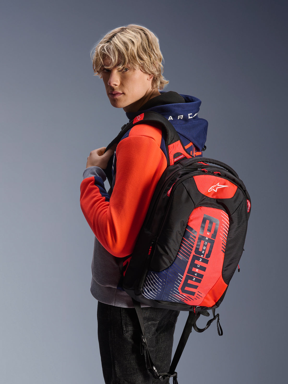 Alpinestars MM93 City Hunter V2 rygsæk, sort med rød fluo og navyblå detaljer, med Marc Marquez MM93 signaturgrafik, båret sammen med en matchende sportshoodie, slank aerodynamisk profil til motorcykelpendling