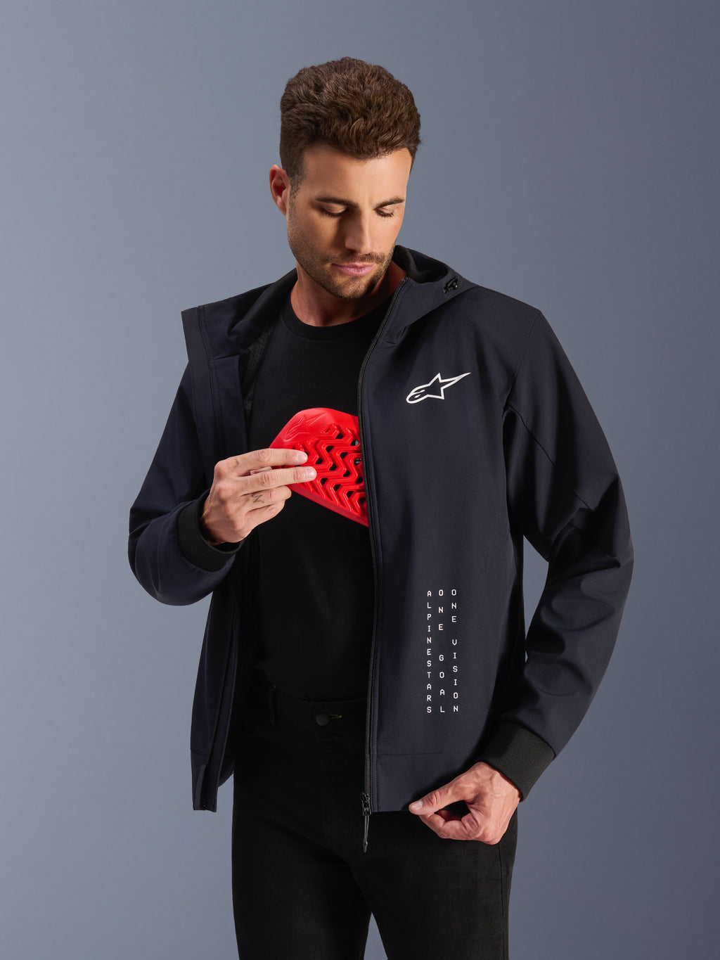 Alpinestars Radium Tech hættetrøje, sort urban motorcykeljakke til herrer, mand holder en rød Nucleon Flex Plus beskyttelsesindsats, CE-certificeret tekstiljakke med hvidt logo, Tech-Air® ready.