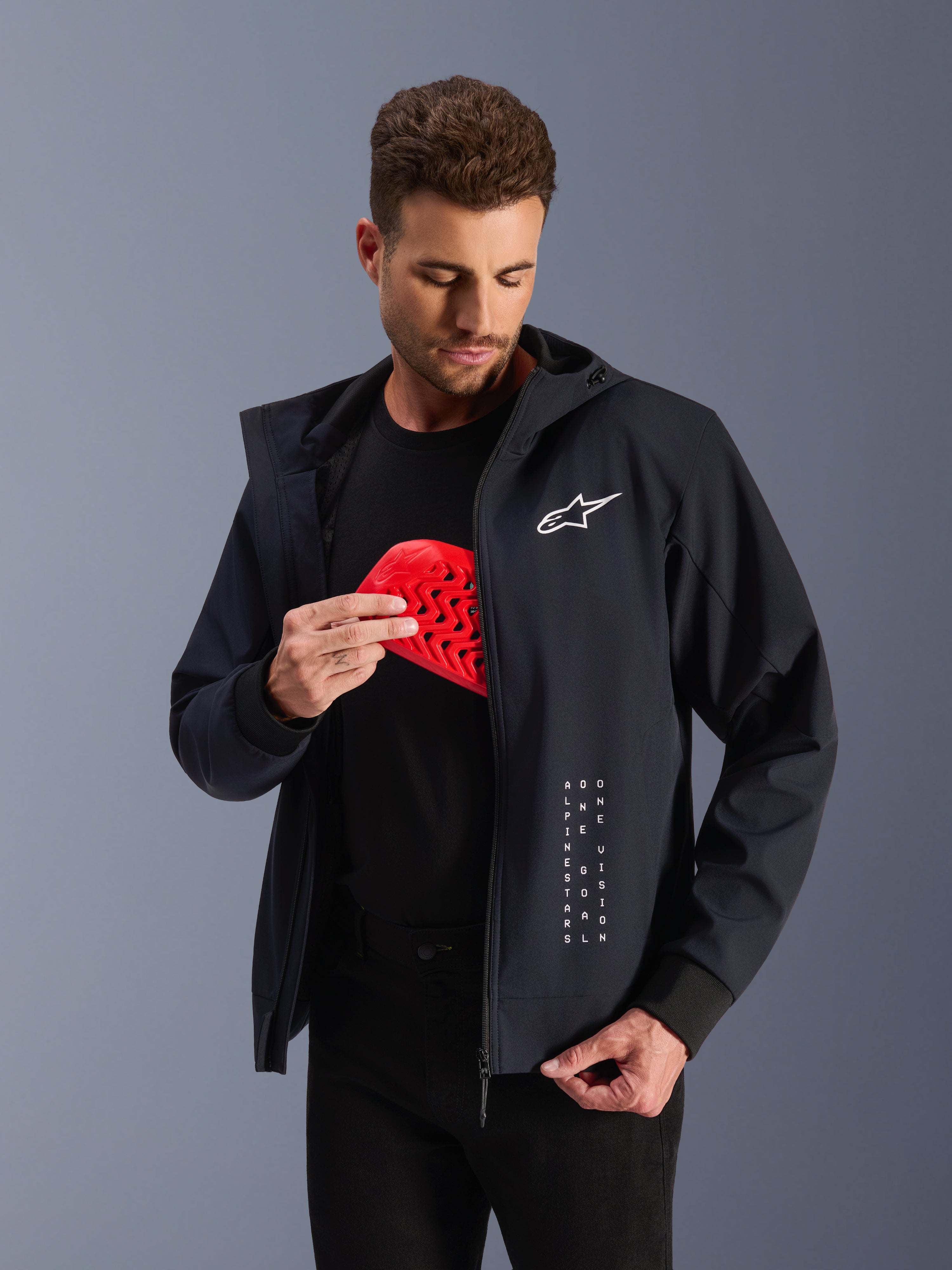 Alpinestars Radium Tech hættetrøje, sort urban motorcykeljakke til herrer, mand holder en rød Nucleon Flex Plus beskyttelsesindsats, CE-certificeret tekstiljakke med hvidt logo, Tech-Air® ready.