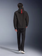 Alpinestars Radium Tech Hoodie, urban motorcykeljakke, sort, set bagfra med klar rød 'ALPINE'-tekst på hætten, kombineret med sorte bukser og sneakers, strømlinet teknisk skal til pendling.
