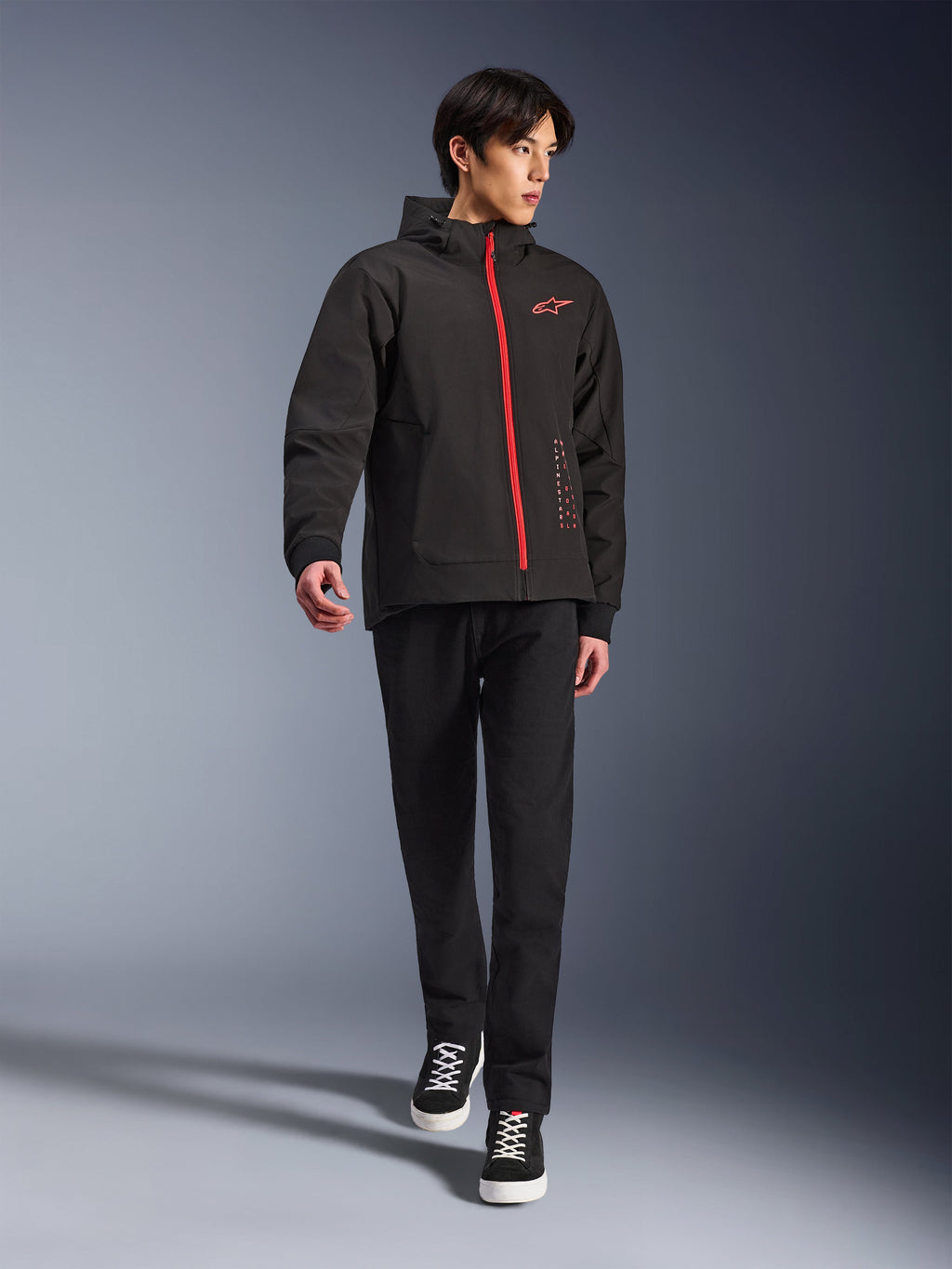 Alpinestars Radium Tech Hoodie i Black Bright Red, urban motorcykeljakke båret af en model, med rød lynlås foran og logo på brystet, stylet med sorte bukser og sneakers for et moderne pendlingslook.