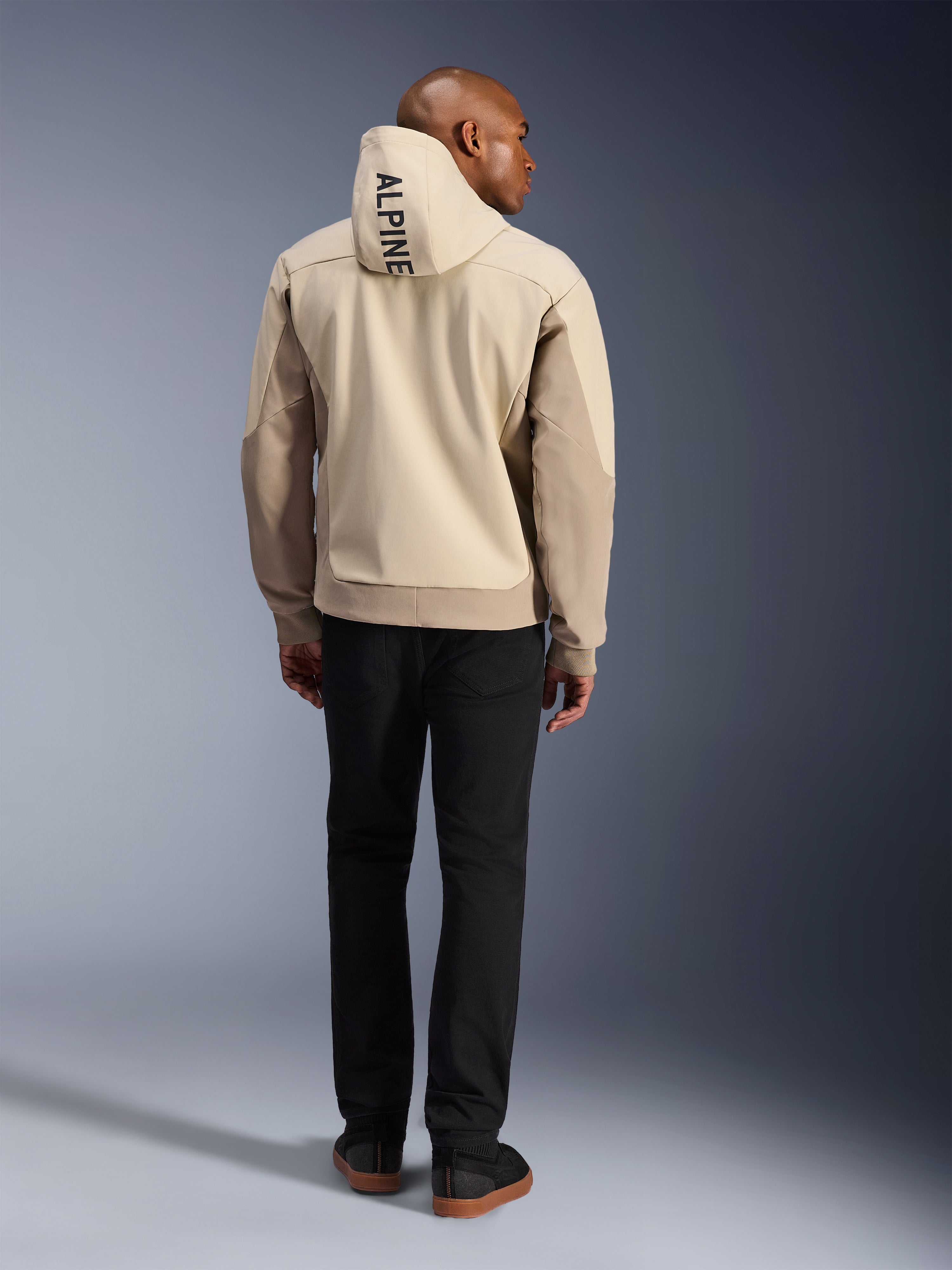 Alpinestars Radium Tech Hoodie, Mastic Aluminum, set bagfra med lodret 'ALPINE'-tekst på hætten, teknisk 3L vindtæt shell urban motorcykeljakke med sandfarvede paneler, båret med sorte bukser