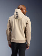 Alpinestars Radium Tech Hoodie, set bagfra af en mand iført den tekniske Mastic Aluminum hoodie, med lodret ALPINE-tekst på hætten, lysebeige og tanfarvede paneler, ribbede manchetter, urban motorcykeljakke