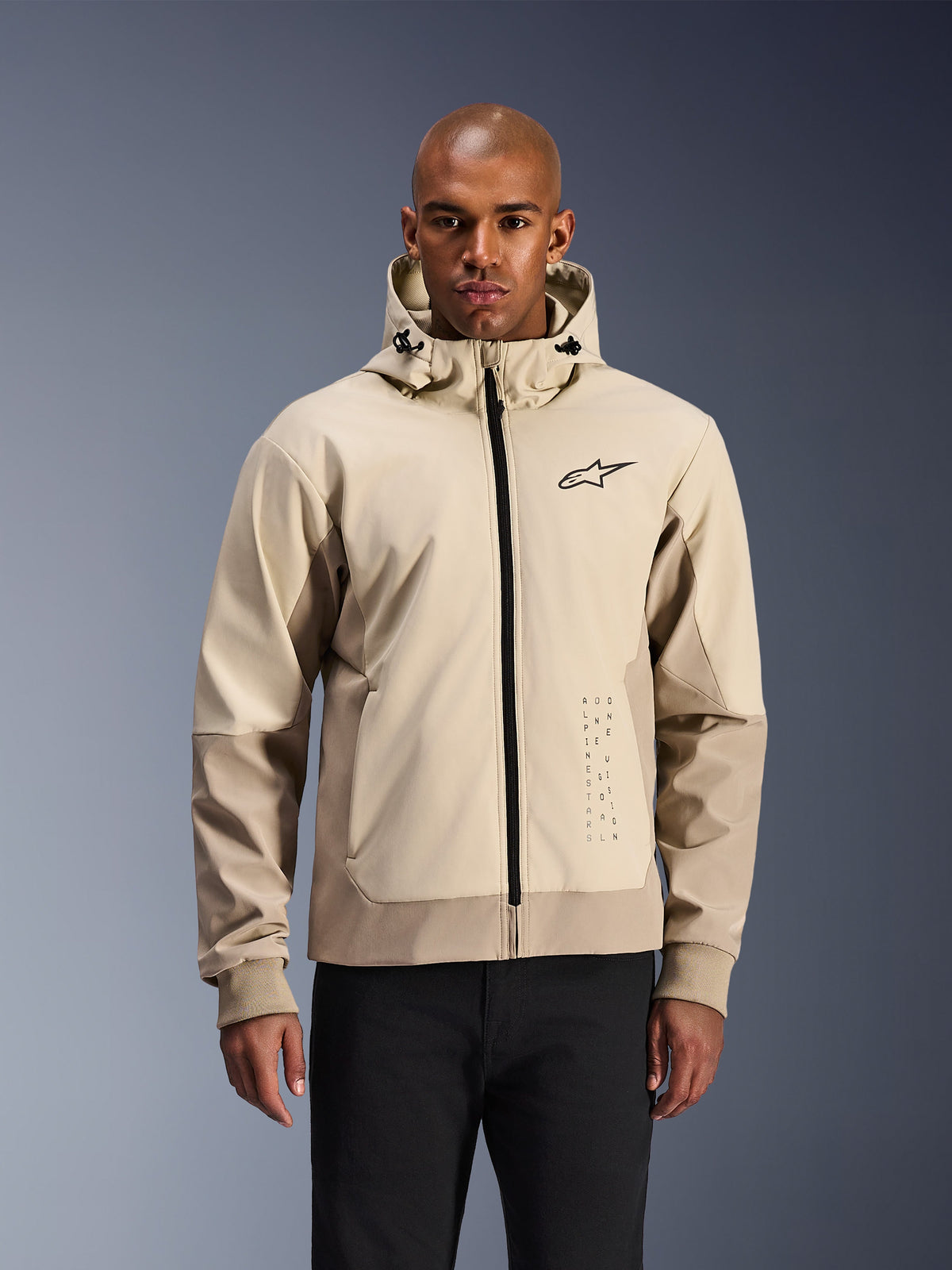 Alpinestars Radium Tech Hoodie in Mastic Aluminum, light beige and tan urban motorcycle jacket, black front zipper and small chest logo, modeled by a man in black pants, technical windproof shell with contemporary streetwear styling