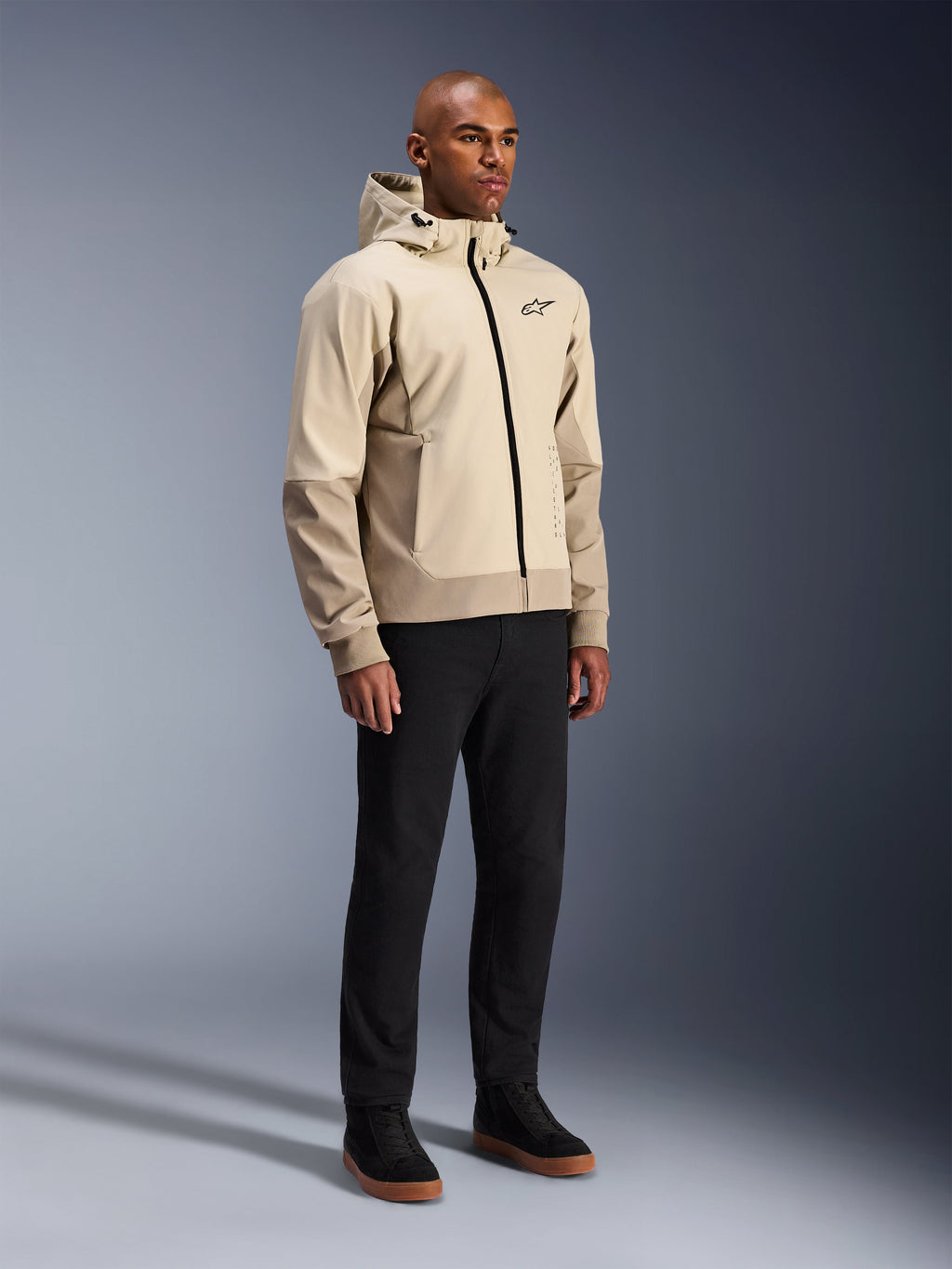 Alpinestars Radium Tech Hoodie, urban motorcykeljakke, Mastic Aluminum lysebeige, båret med sorte bukser og sorte high-top sneakers, med en teknisk vindtæt skal, lynlås foran og aftagelig hætte, moderne streetwear-design til bykørsel