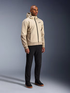 Alpinestars Radium Tech Hoodie, urban motorcykeljakke, Mastic Aluminum lysebeige, båret med sorte bukser og sorte high-top sneakers, med en teknisk vindtæt skal, lynlås foran og aftagelig hætte, moderne streetwear-design til bykørsel