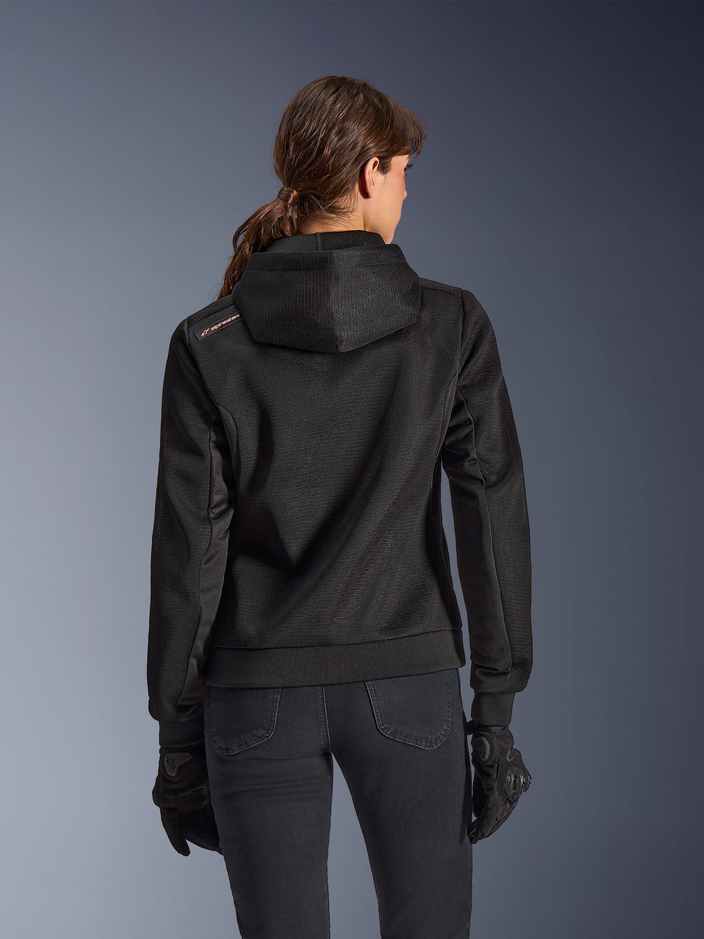 Alpinestars Stella Chrome Superairflow Hoodie, set bagfra af kvinde iført sort teknisk 3D-mesh motorcykelhættetrøje, har konstrueret hætte og kobberfarvet logo på skulderen, kombineret med sorte motorcykelhandsker og bukser.