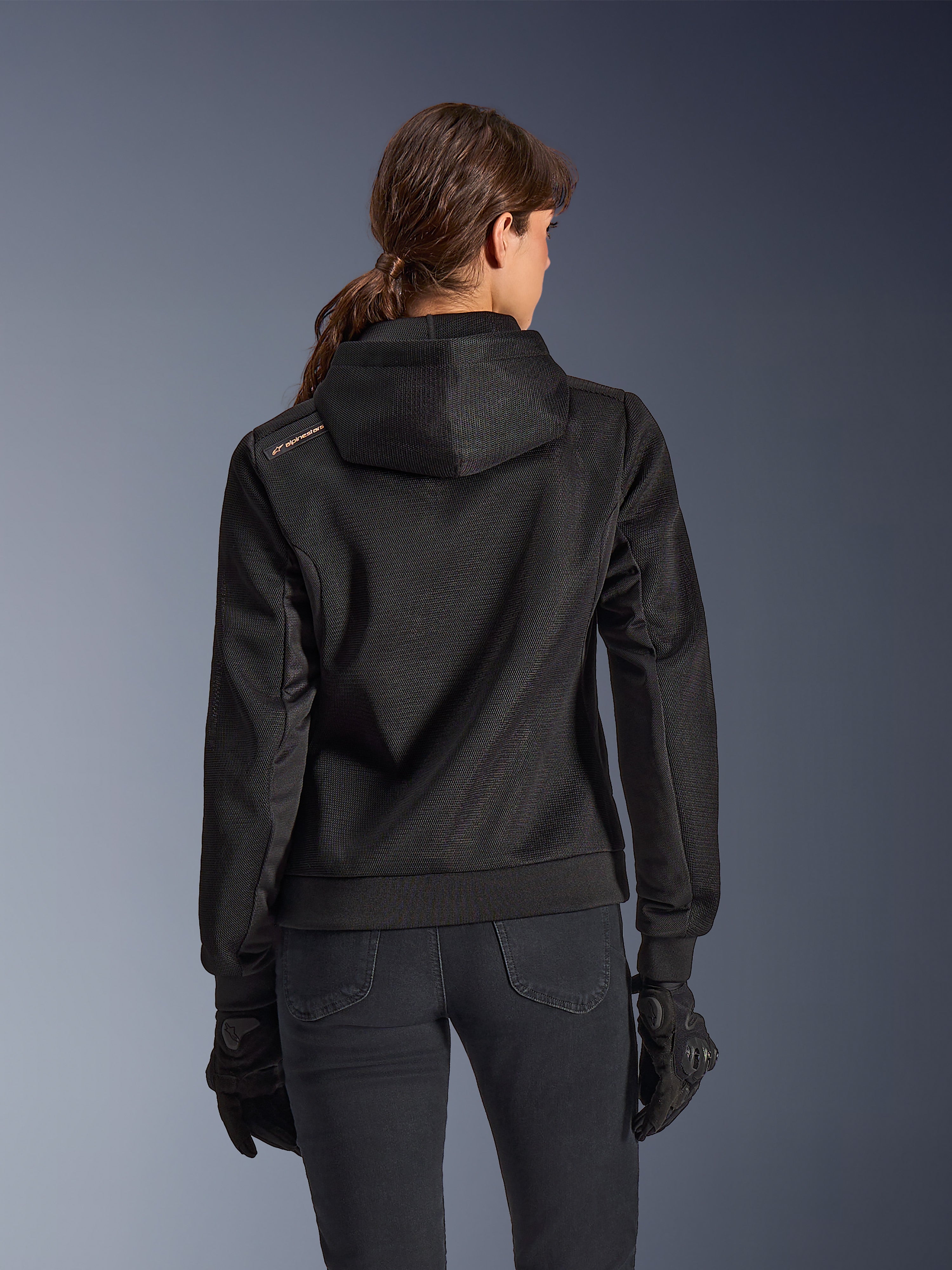 Alpinestars Stella Chrome Superairflow Hoodie, set bagfra af kvinde iført sort teknisk 3D-mesh motorcykelhættetrøje, har konstrueret hætte og kobberfarvet logo på skulderen, kombineret med sorte motorcykelhandsker og bukser.