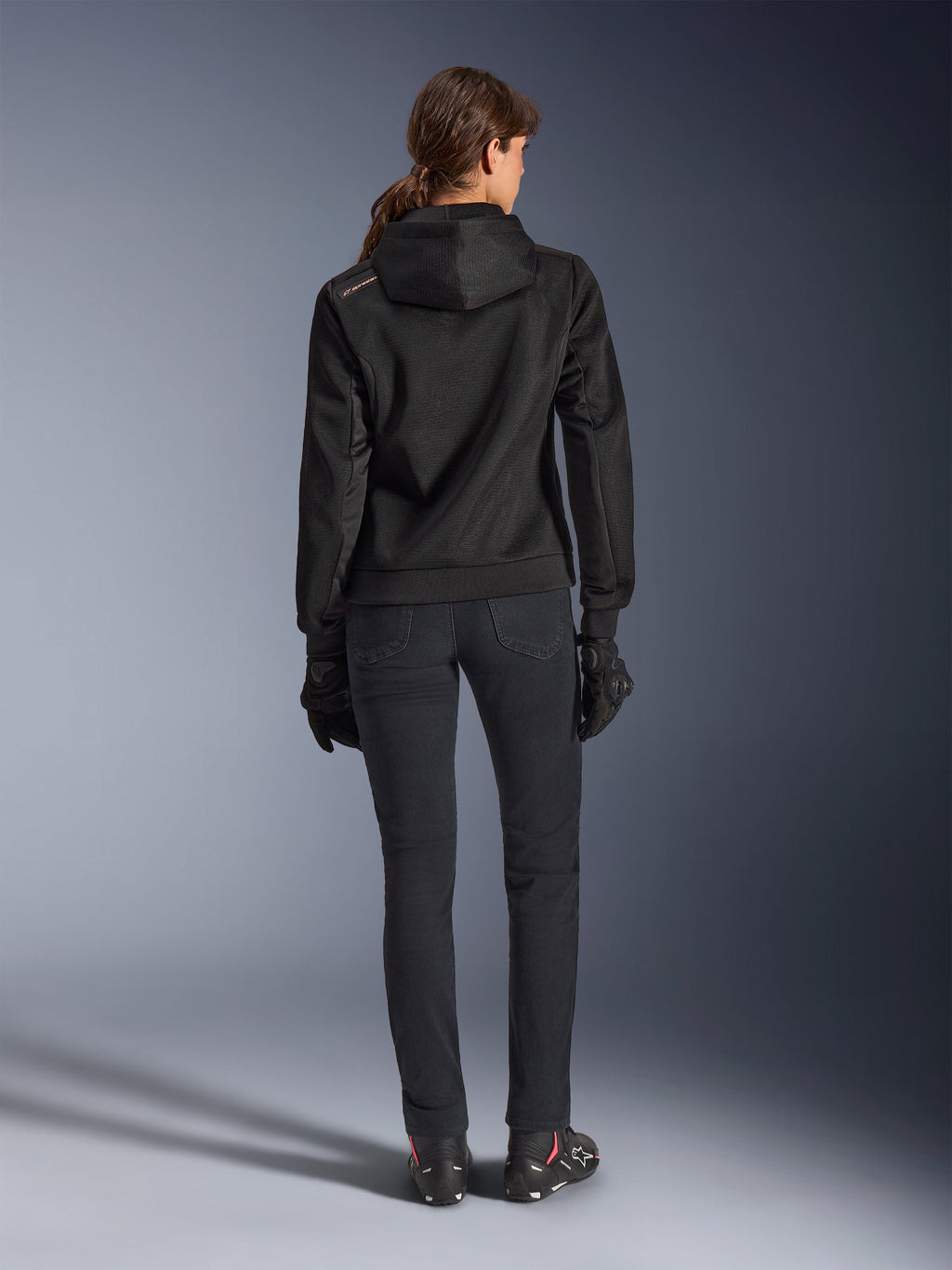 Alpinestars Stella Chrome Superairflow Hoodie, teknisk motorcykelhættetrøje til kvinder, sort 3D-mesh med lille orange logo på skulderen, set bagfra, båret med sorte kørebukser, sorte handsker og støvler, designet til urban sportskørsel