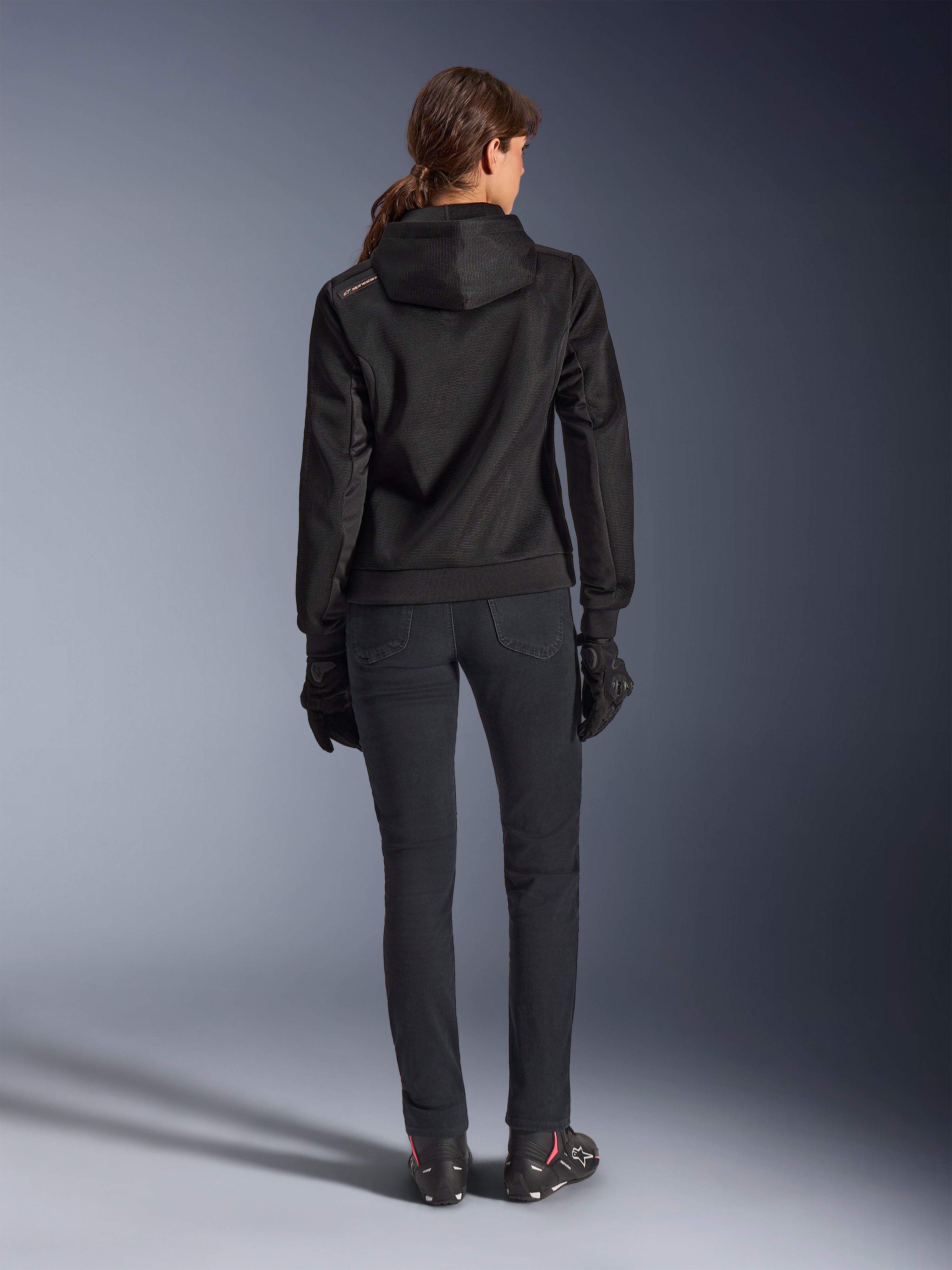 Alpinestars Stella Chrome Superairflow Hoodie, teknisk motorcykelhættetrøje til kvinder, sort 3D-mesh med lille orange logo på skulderen, set bagfra, båret med sorte kørebukser, sorte handsker og støvler, designet til urban sportskørsel