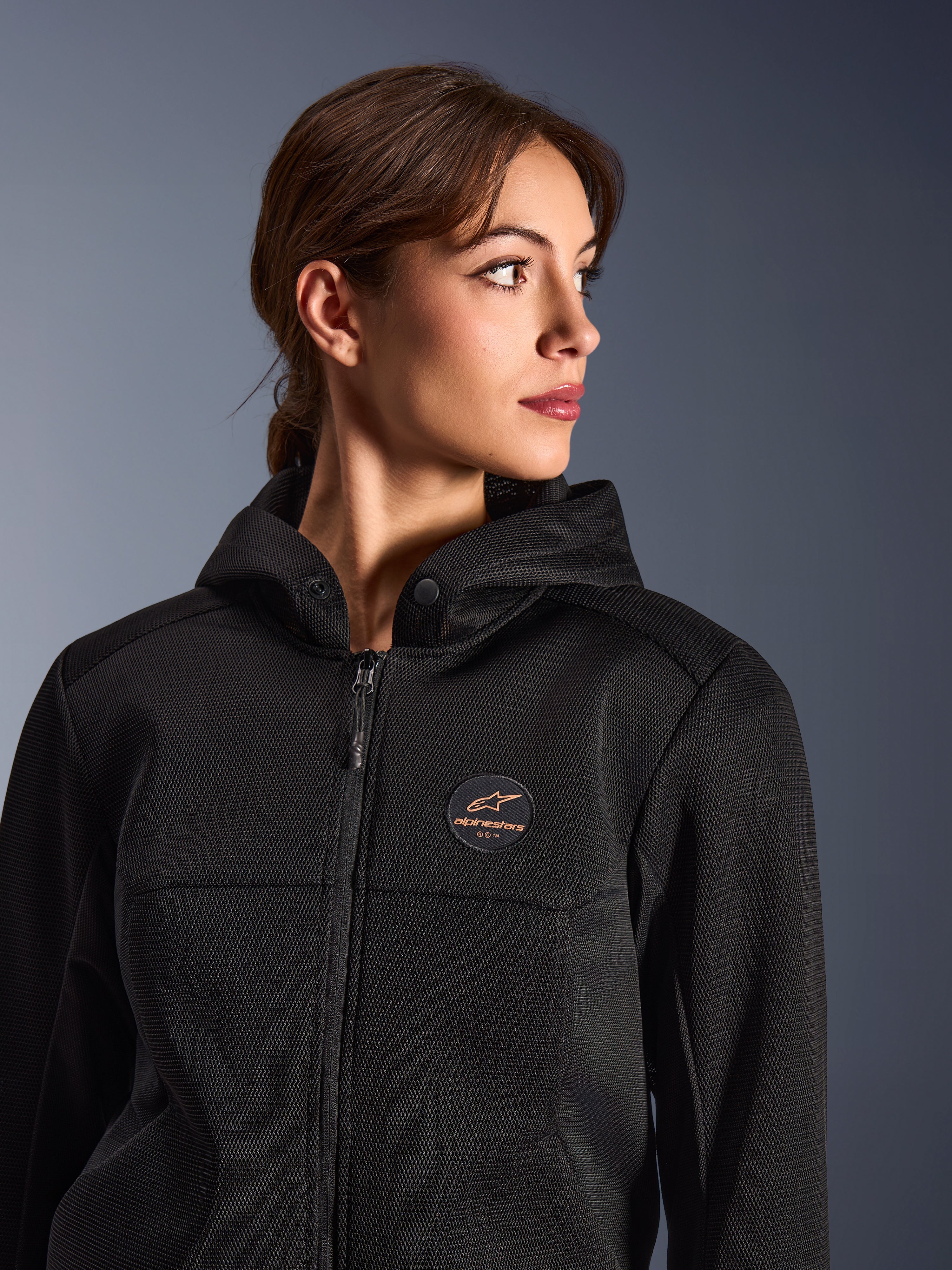 Kvinde iført Alpinestars Stella Chrome Superairflow hættetrøje, sort med kobberfarvet logo, 3D-mesh-konstruktion for maksimal luftgennemstrømning, teknisk motorcykeludstyr designet til kvinder, urban sport-kørestil